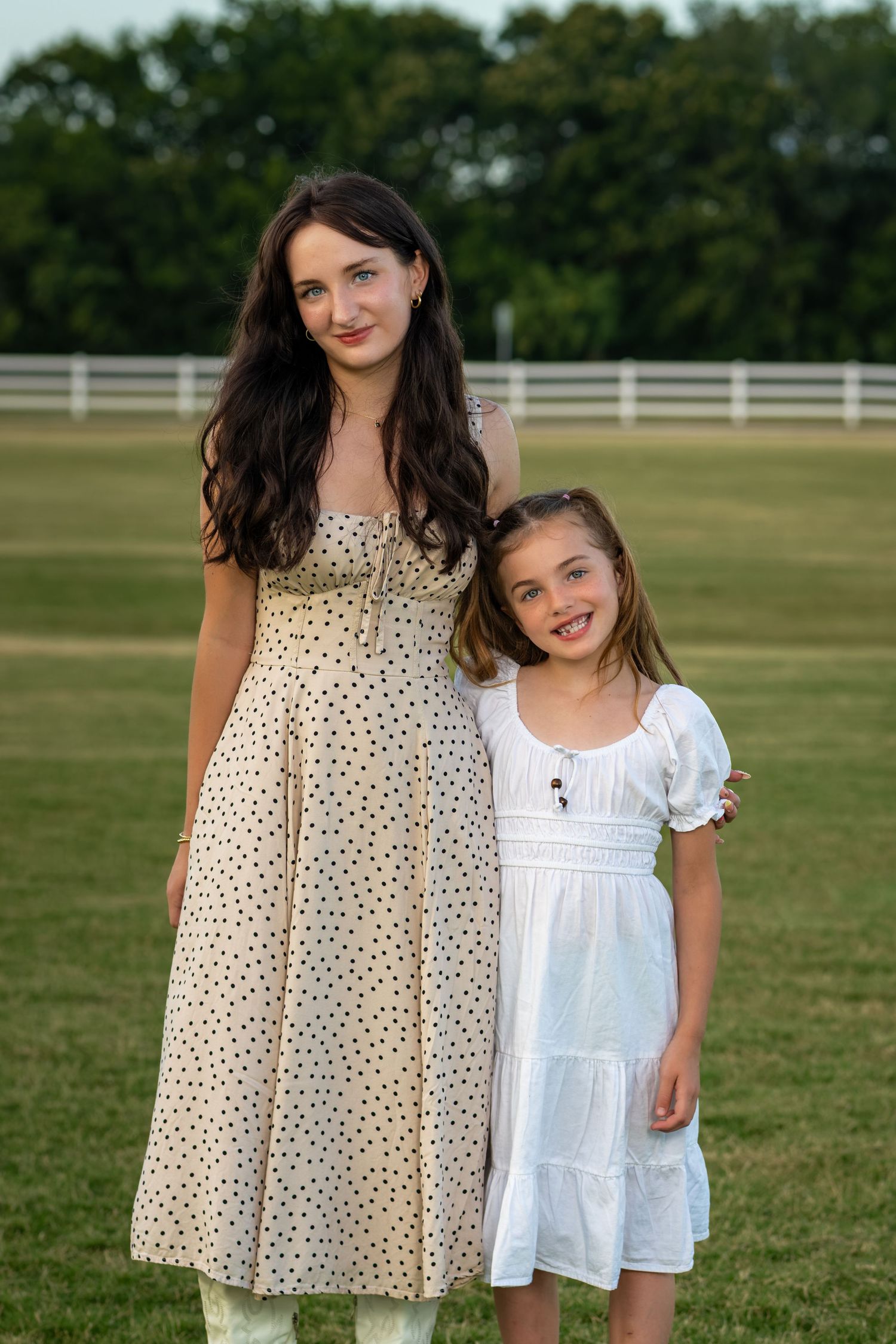 A woman in a polka dot dress and a young girl in white dress pose together outdoors on a green lawn.