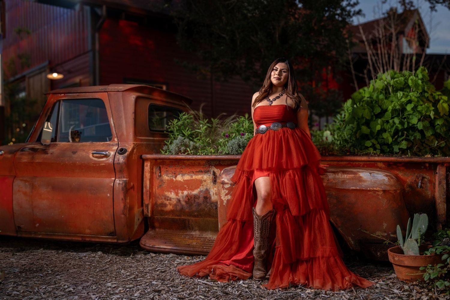 Woman in red tulle gown and cowboy boots poses beside a rustic vintage red pickup truck outdoors.