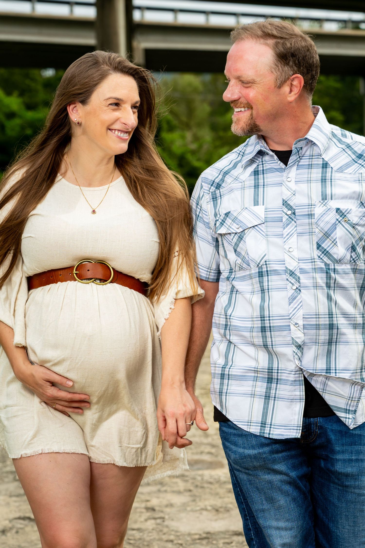 A smiling pregnant woman in a cream dress holds hands with a man in a plaid shirt outdoors near a bridge.