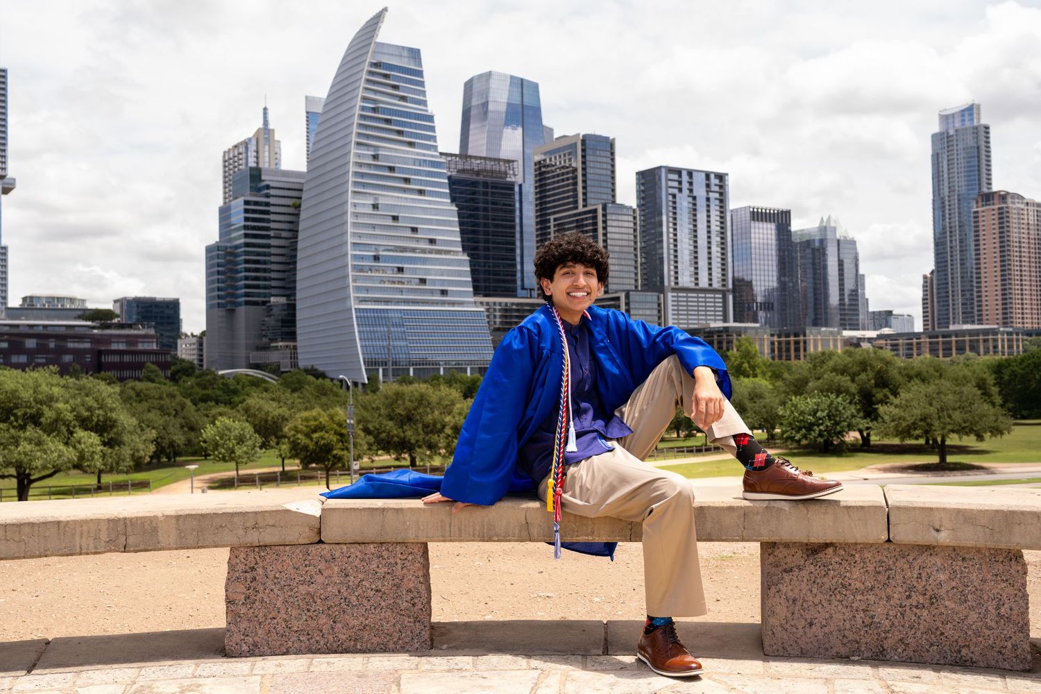 Young man in blue graduation gown sits on stone wall with Dallas city skyline in background.