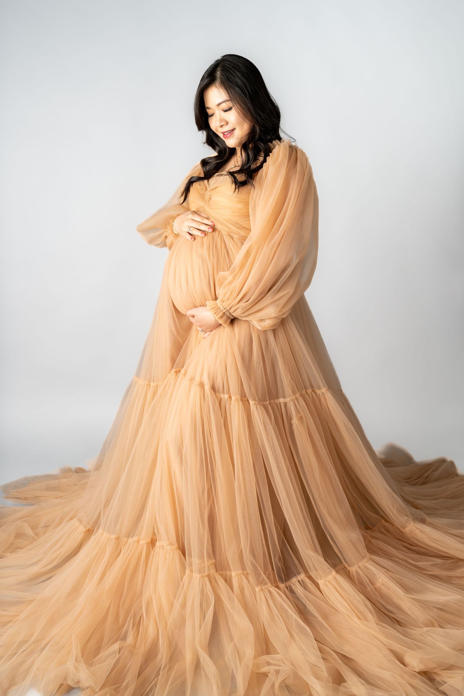Pregnant woman in elegant peach tulle maternity gown cradling baby bump in professional studio photo.