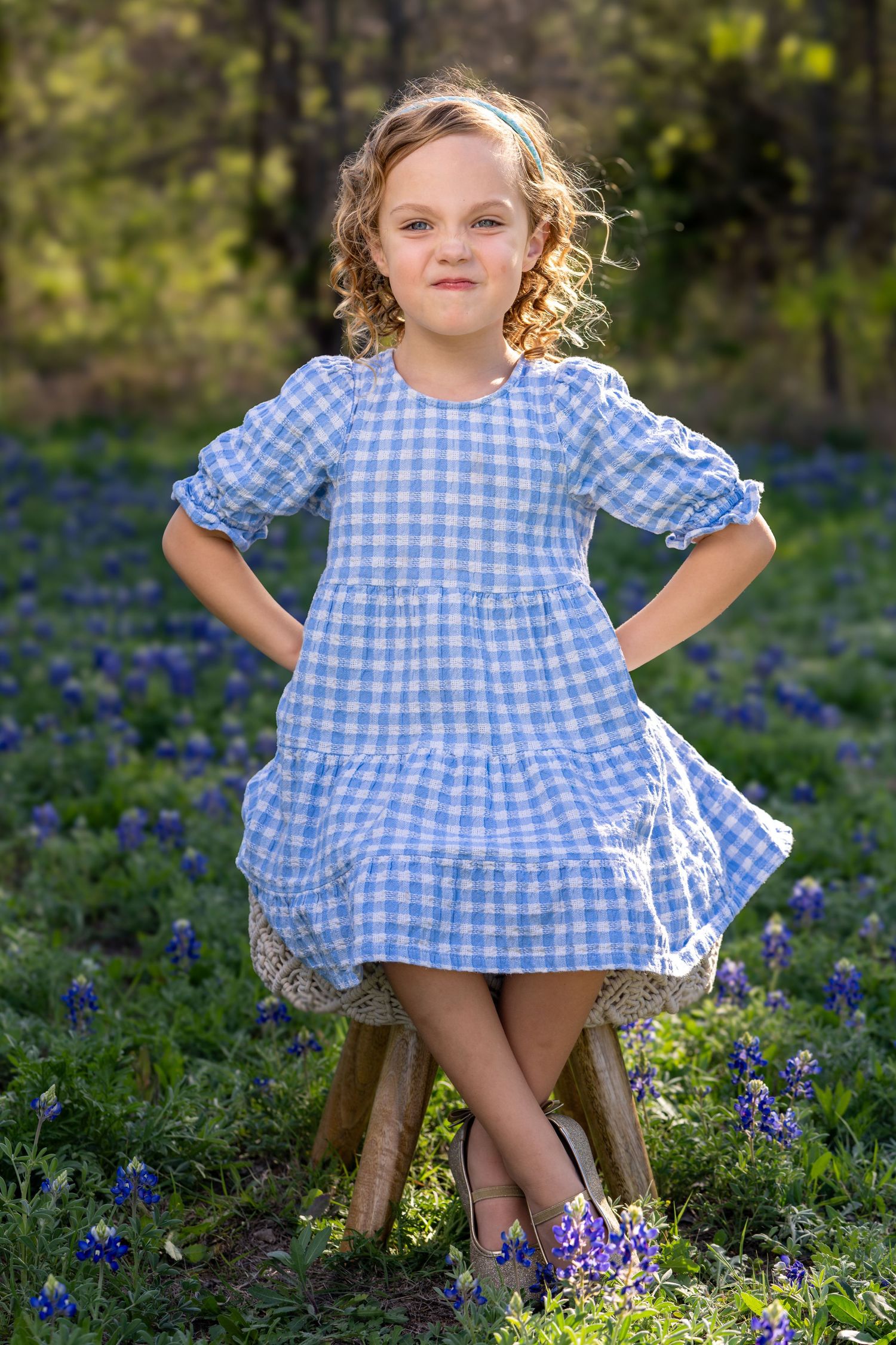 A child in a blue gingham dress poses playfully among blooming bluebonnets in a sunny garden setting.