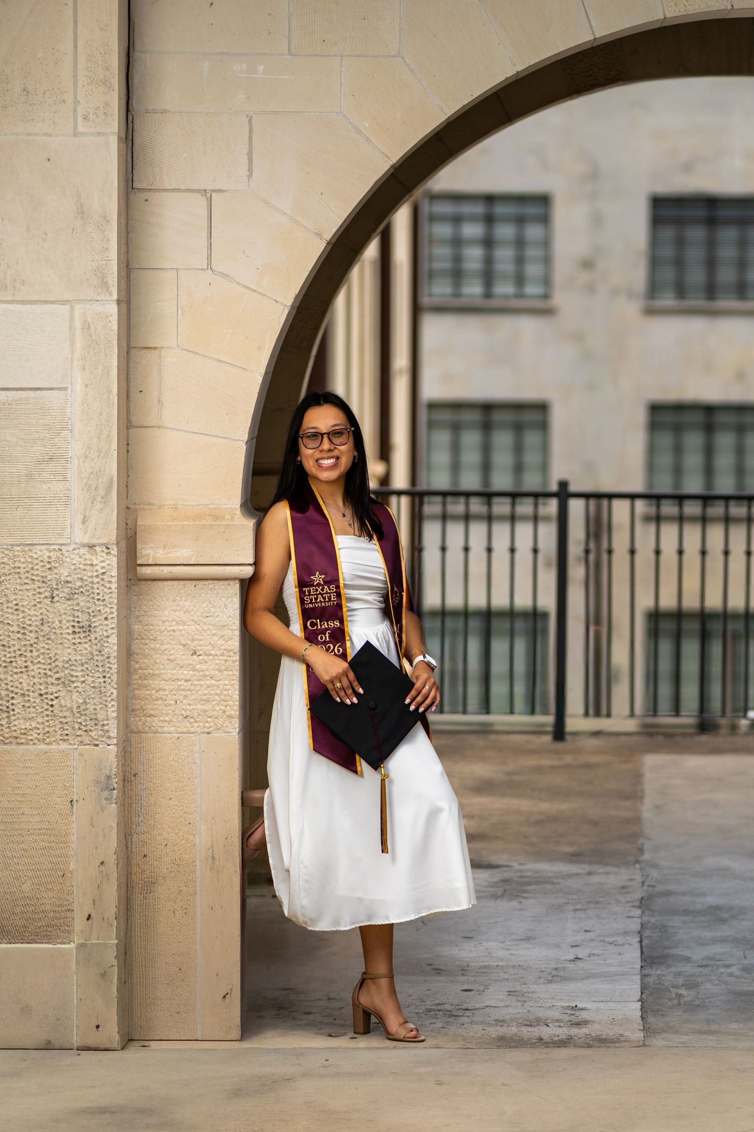 Young woman in white dress and graduation stole holds cap, posing under stone arch at university.