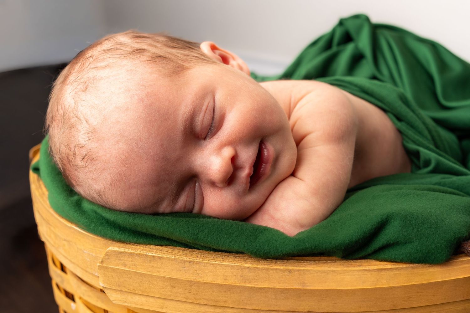 Smiling newborn baby sleeping peacefully in a yellow basket wrapped in a soft green blanket.