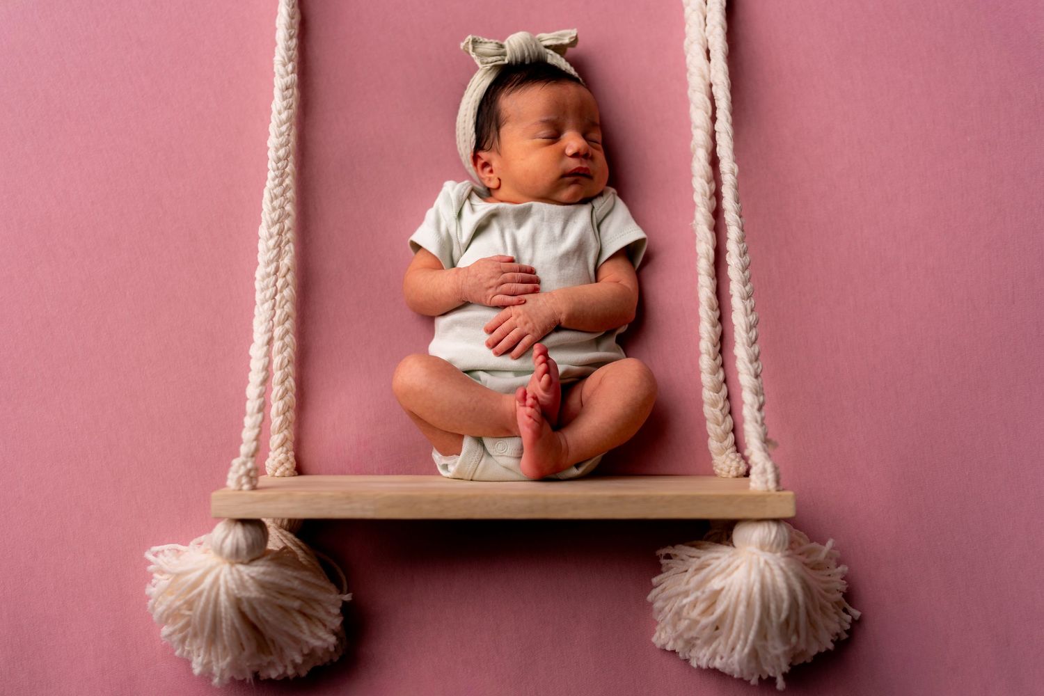A newborn baby sleeping peacefully on a swing prop with a pink background.
