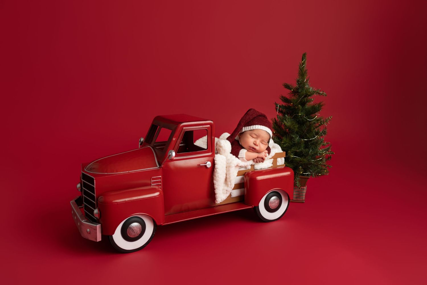 Newborn baby in a Santa hat sleeping in a vintage red toy truck beside a small Christmas tree on a red background.