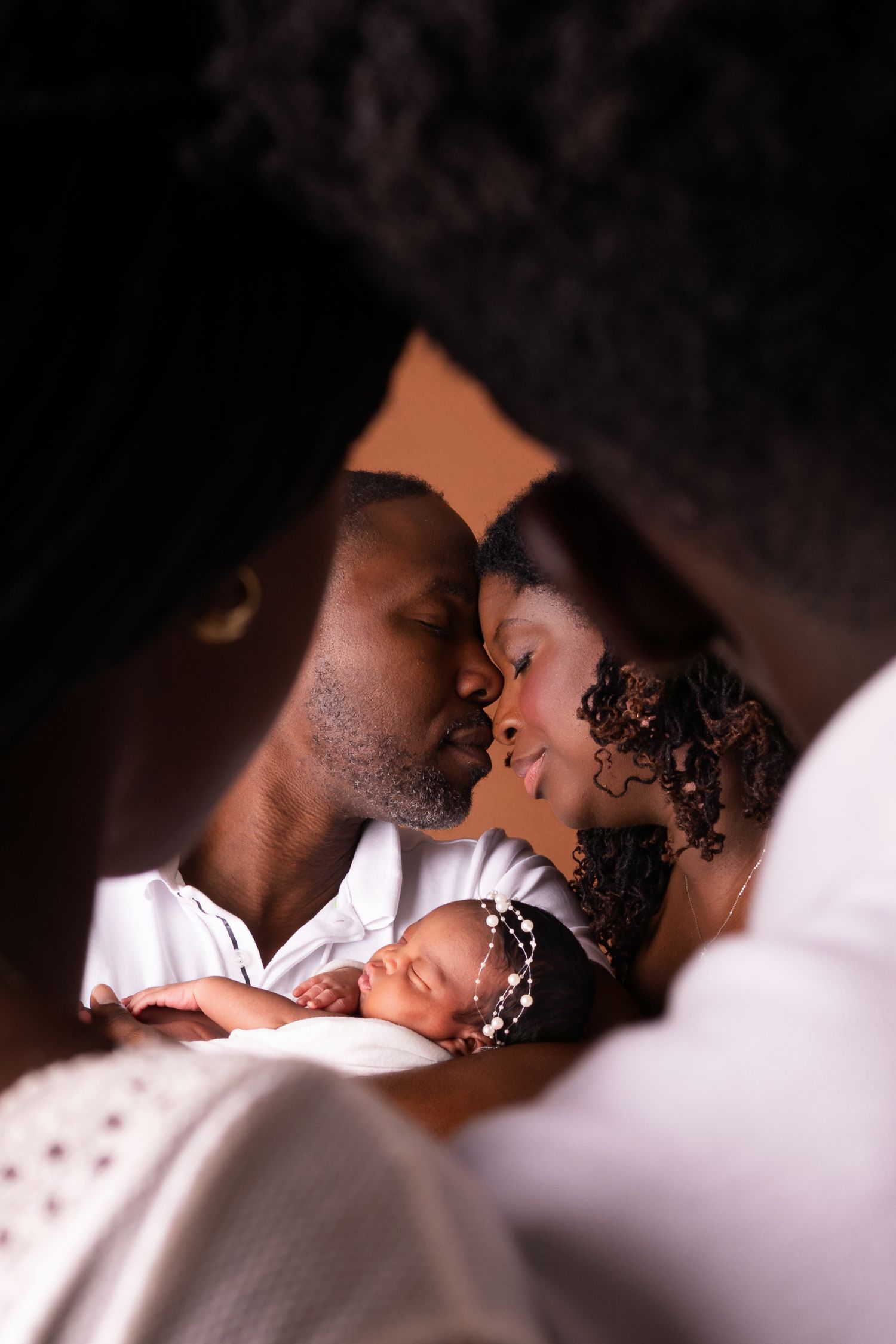 Tender newborn family portrait of two parents touching foreheads as their sleeping baby in a beaded headband rests between them.