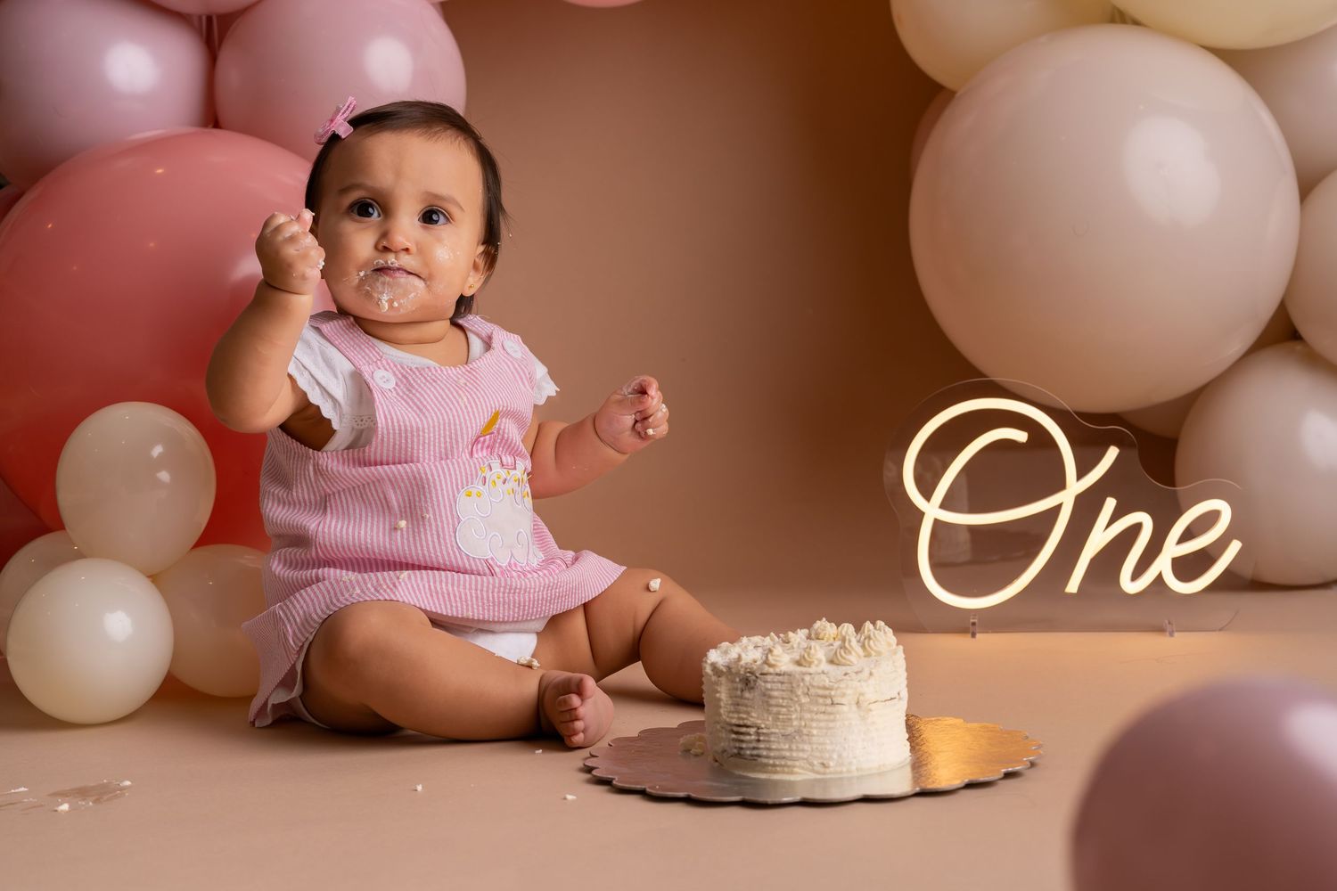Baby girl in pink romper celebrates first birthday smashing cake surrounded by pink and white balloons.