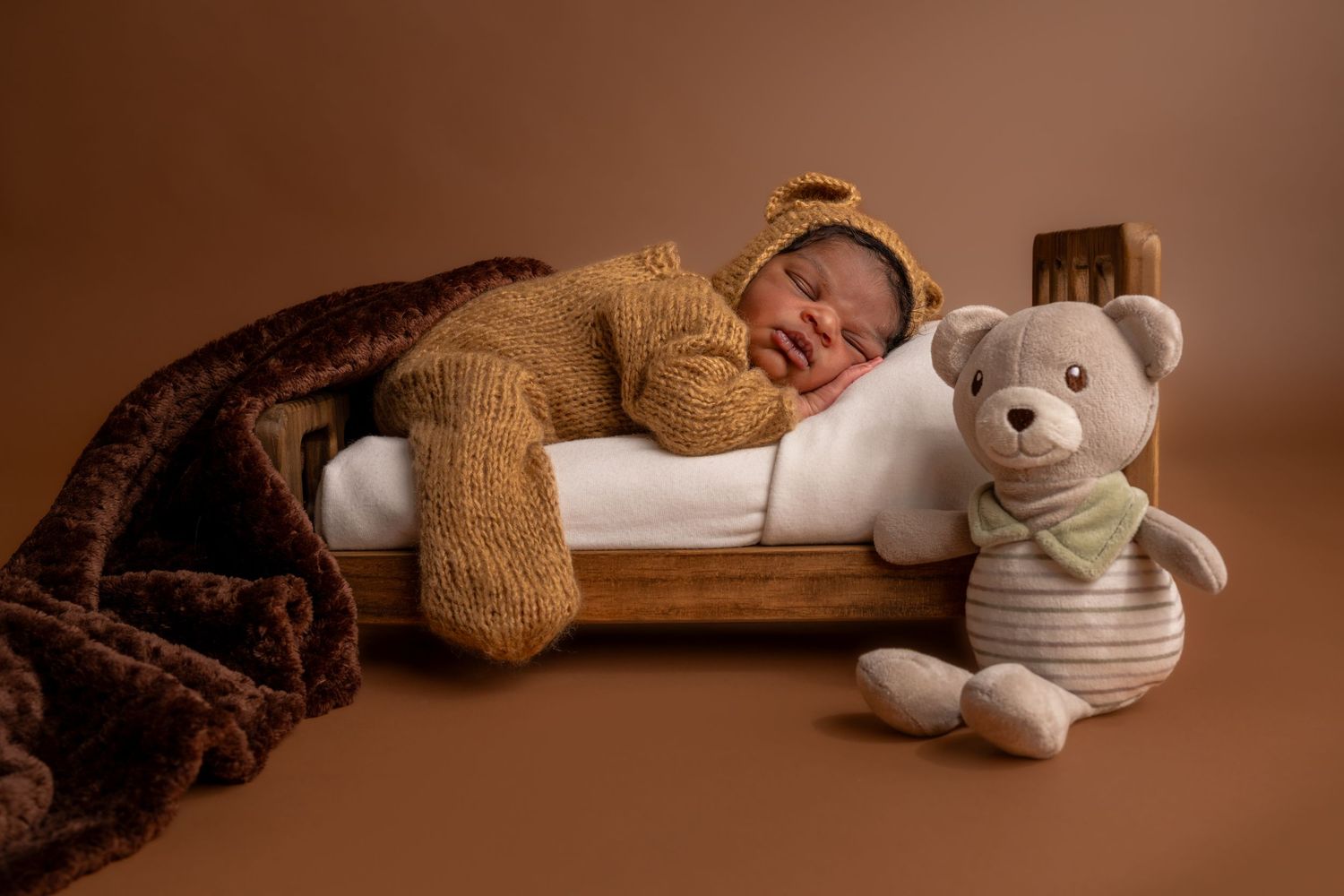 Newborn baby in brown bear knit outfit sleeping on tiny wooden bed beside a teddy bear on brown background.