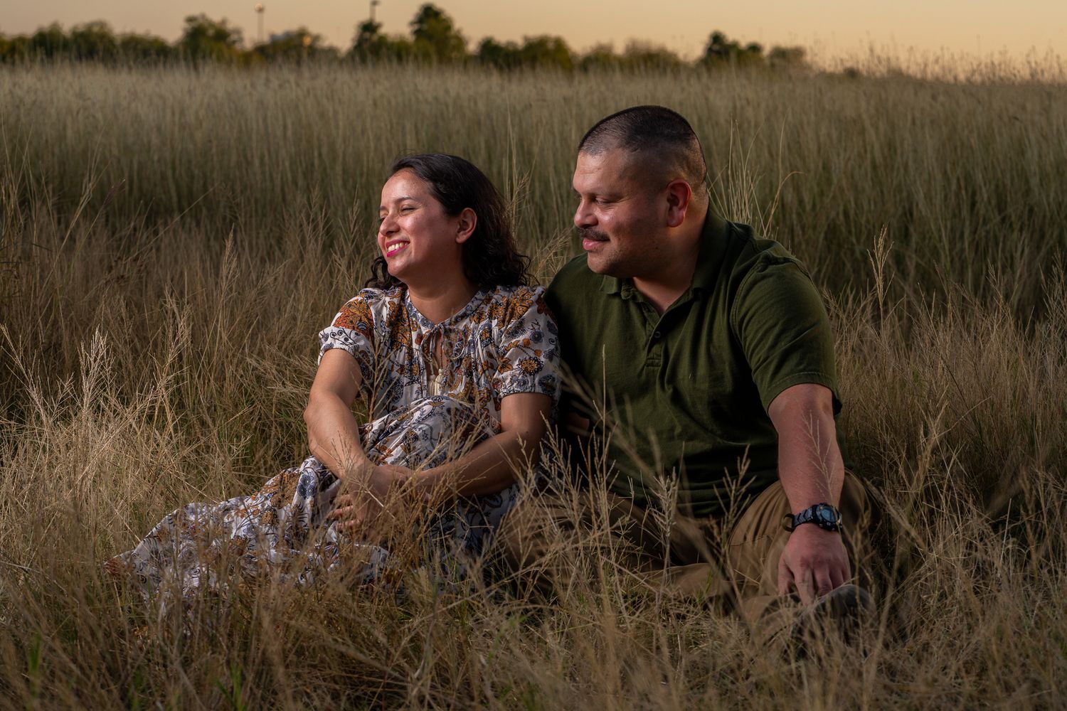 Family photography: A couple sits together laughing in a golden grass field at sunset during a romantic outdoor moment in Texas.