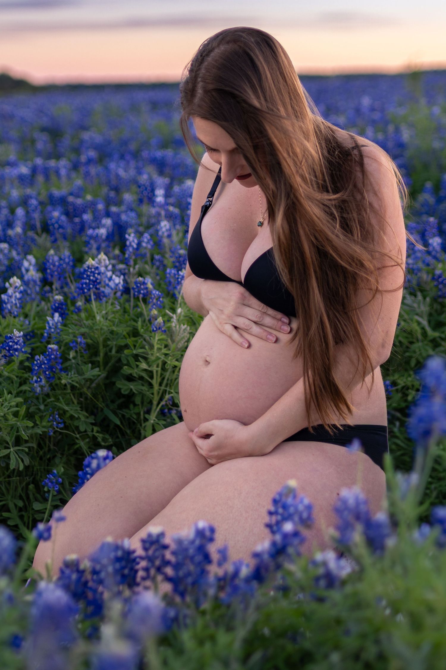 Pregnant woman in black bikini sitting among blooming bluebonnets at sunset, cradling her baby bump.
