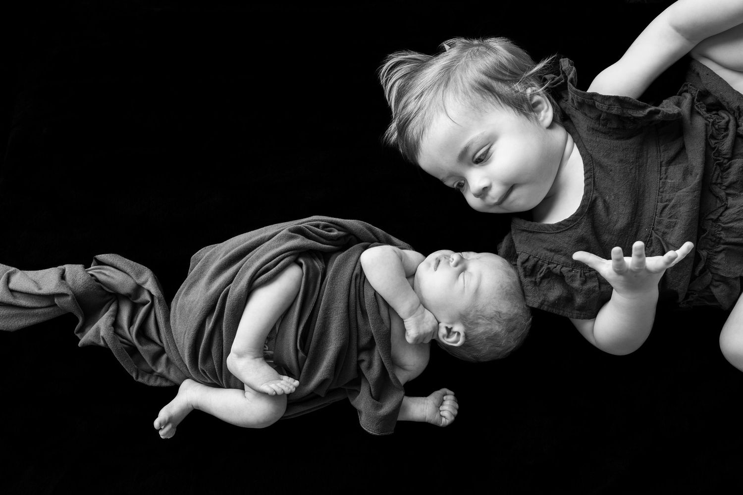 Black and white photo of a toddler leaning over a newborn baby against a dark background.