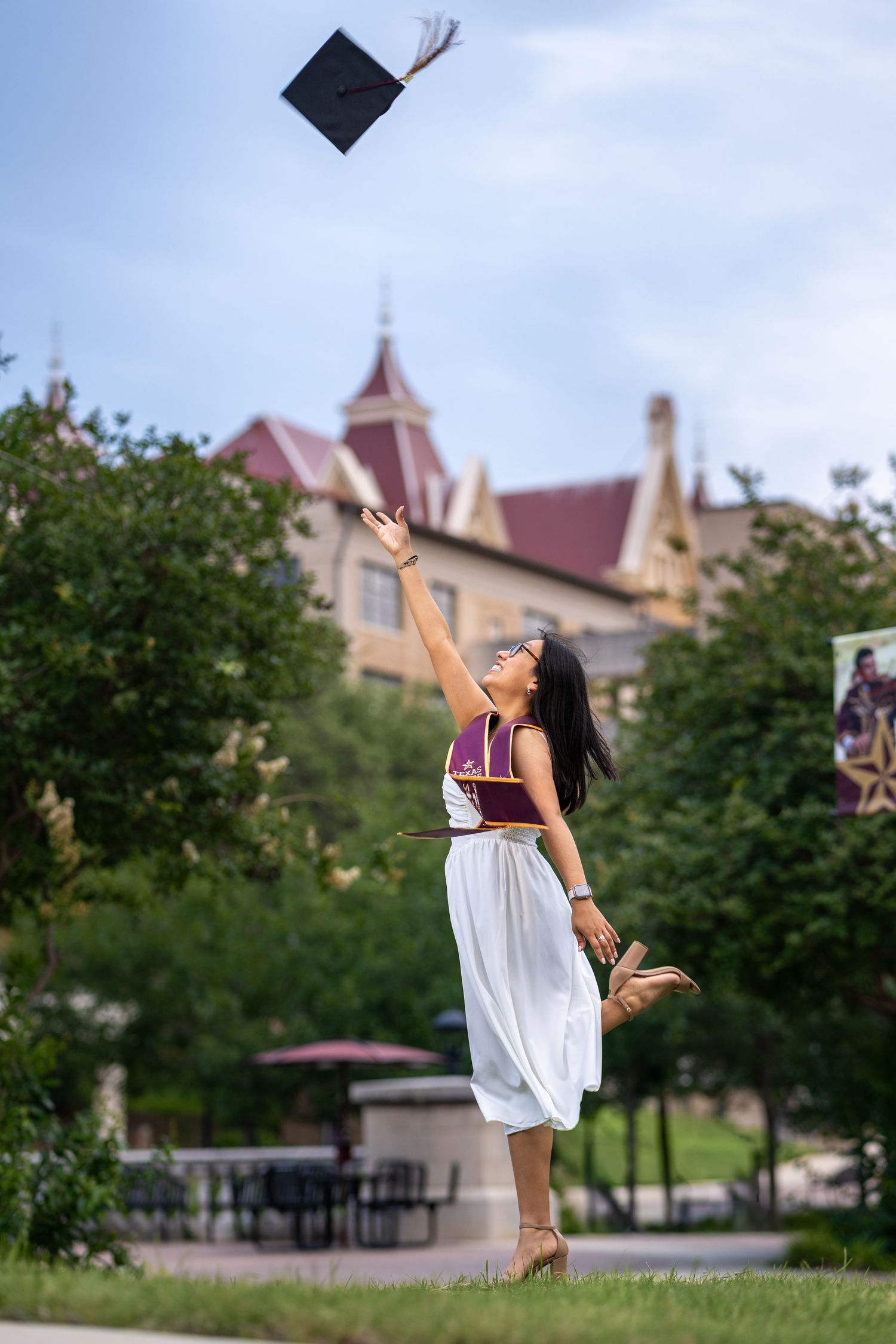 Young woman in white dress joyfully tossing graduation cap in the air on a college campus.