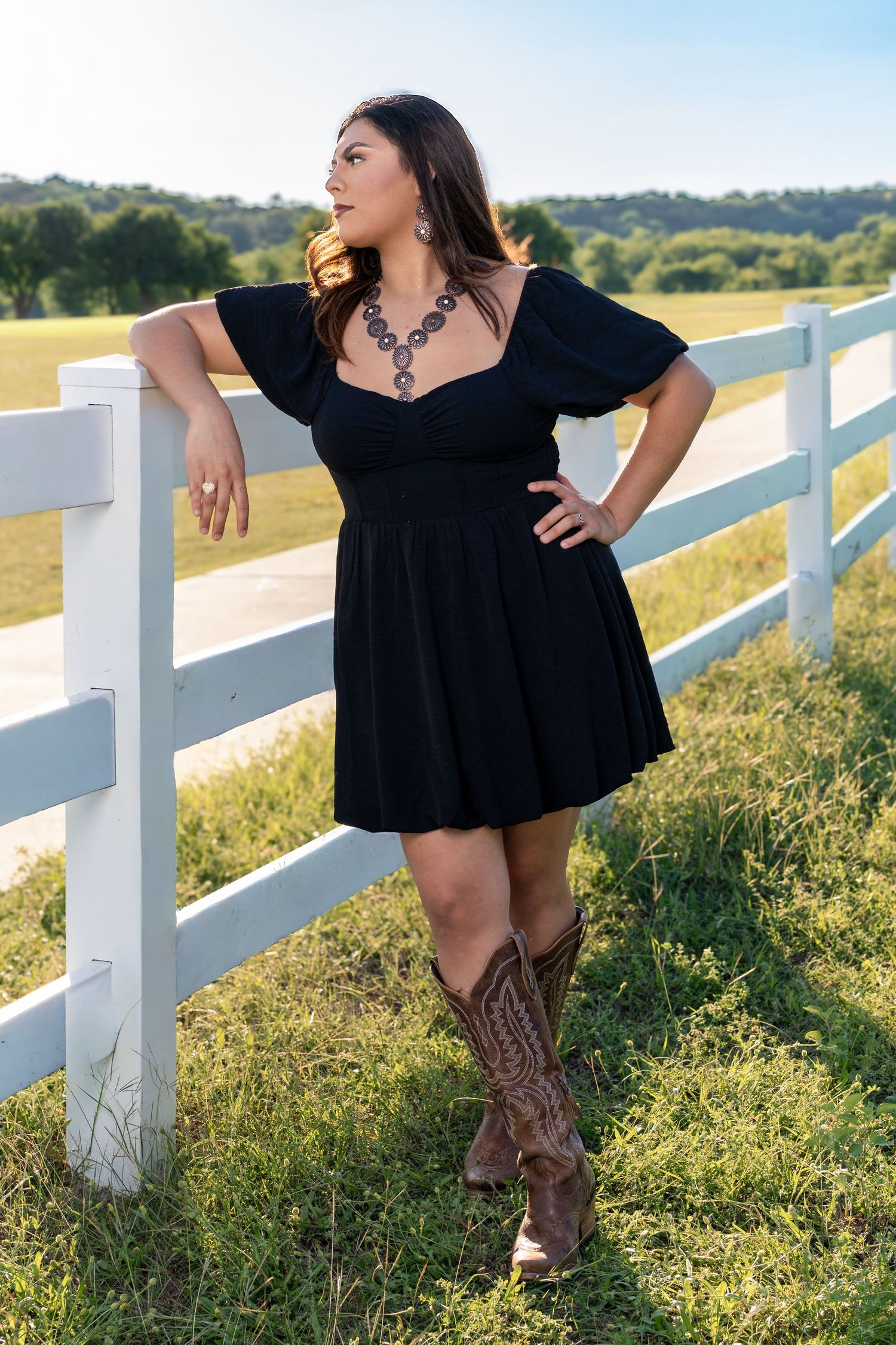 Young woman in black dress and cowboy boots poses by a white fence in a sunny green field.