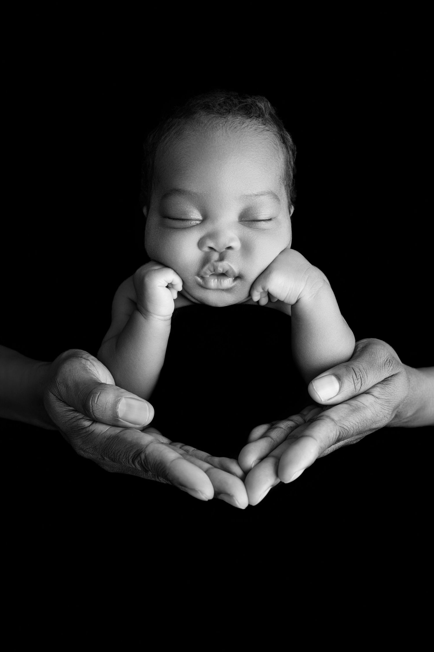 Black and white portrait of a sleeping newborn baby with chin resting on tiny fists, cradled in two elderly hands against a dark background.