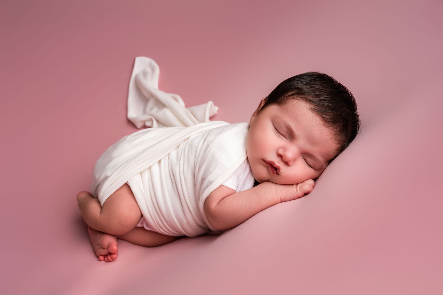 Sleeping newborn baby wrapped in a soft white cloth, lying on its side with hand tucked under cheek against a pink backdrop.