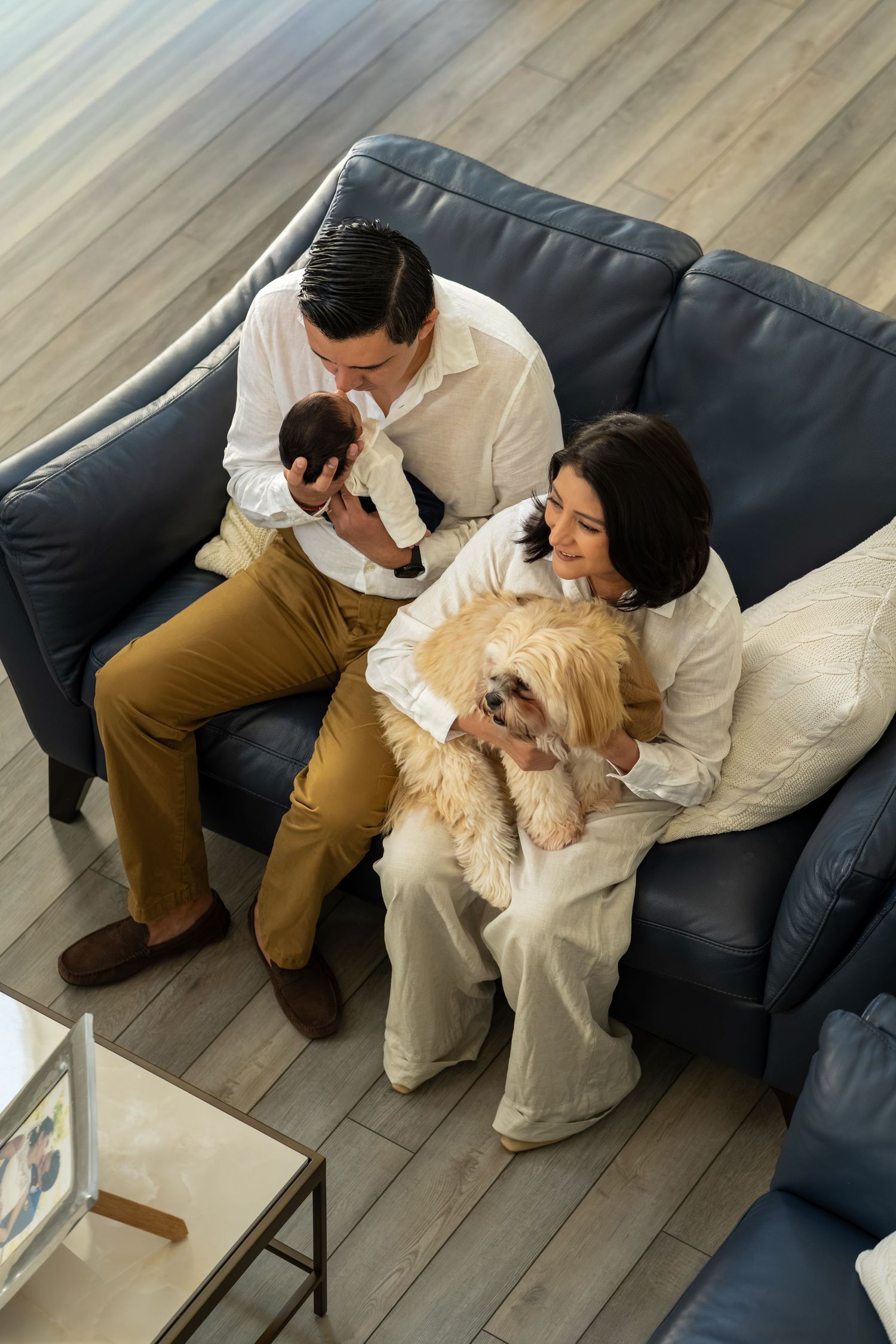 Family sitting on blue leather sofa with newborn baby and fluffy golden dog in modern home.