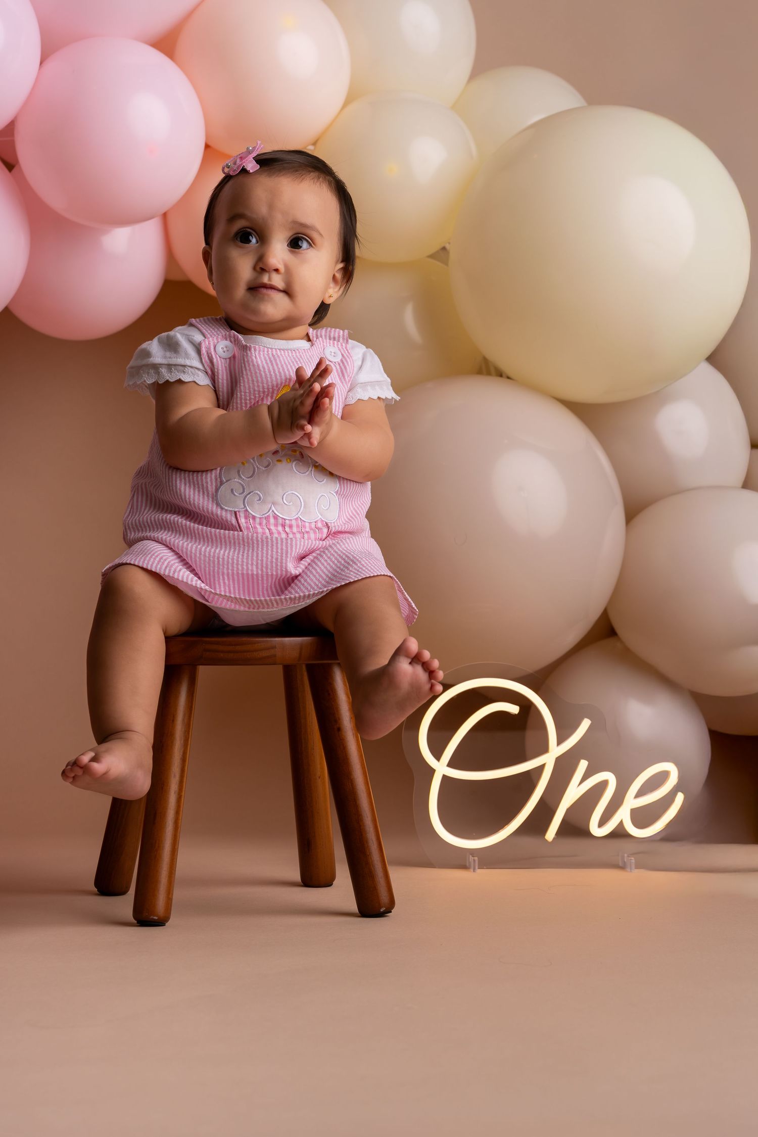 First birthday photo setup with pink and white balloons, wooden stool, and glowing 'One' neon sign against beige backdrop.