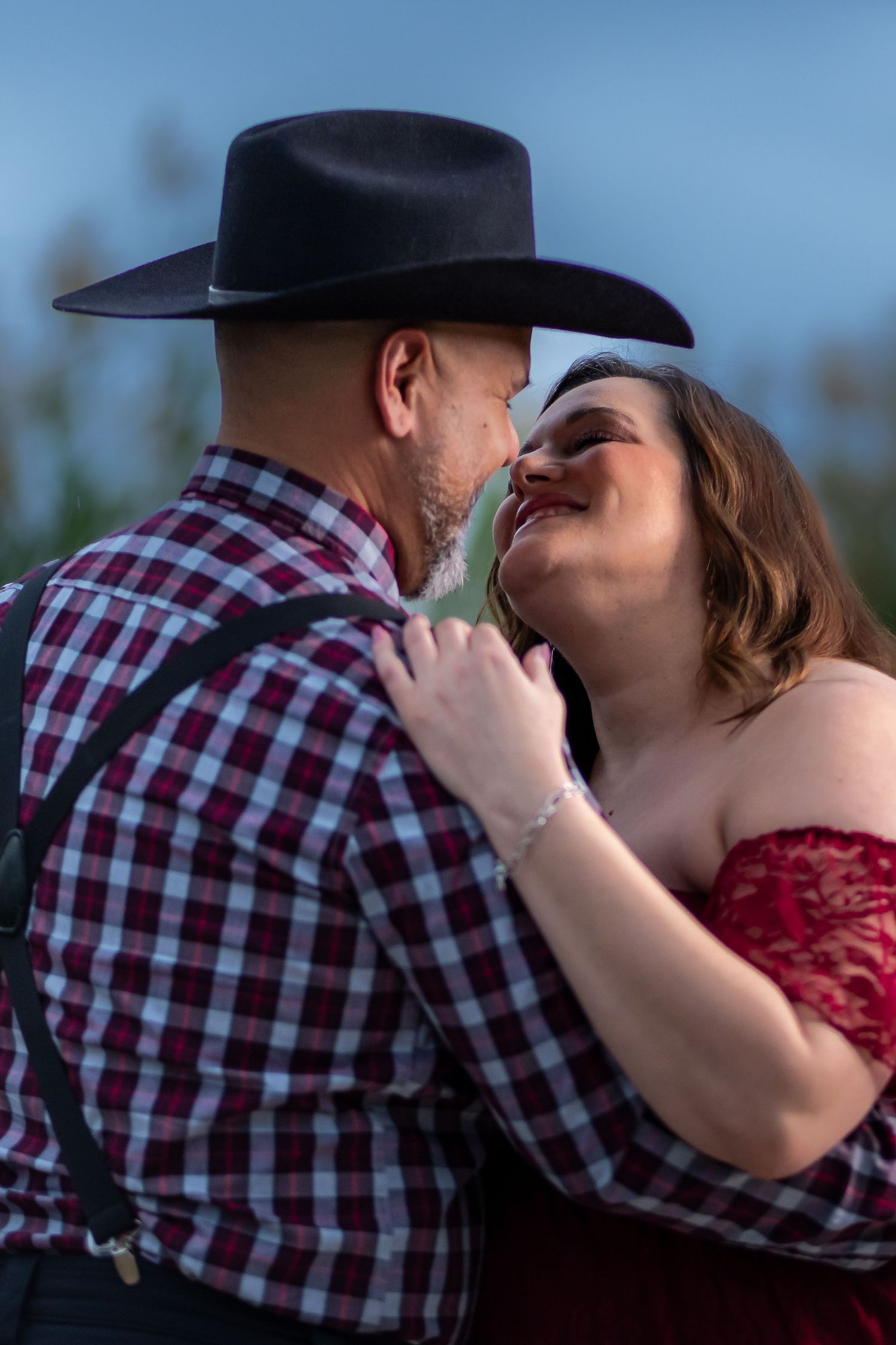 A romantic couple embraces outdoors, with one wearing a cowboy hat and plaid shirt and the other in a red dress.