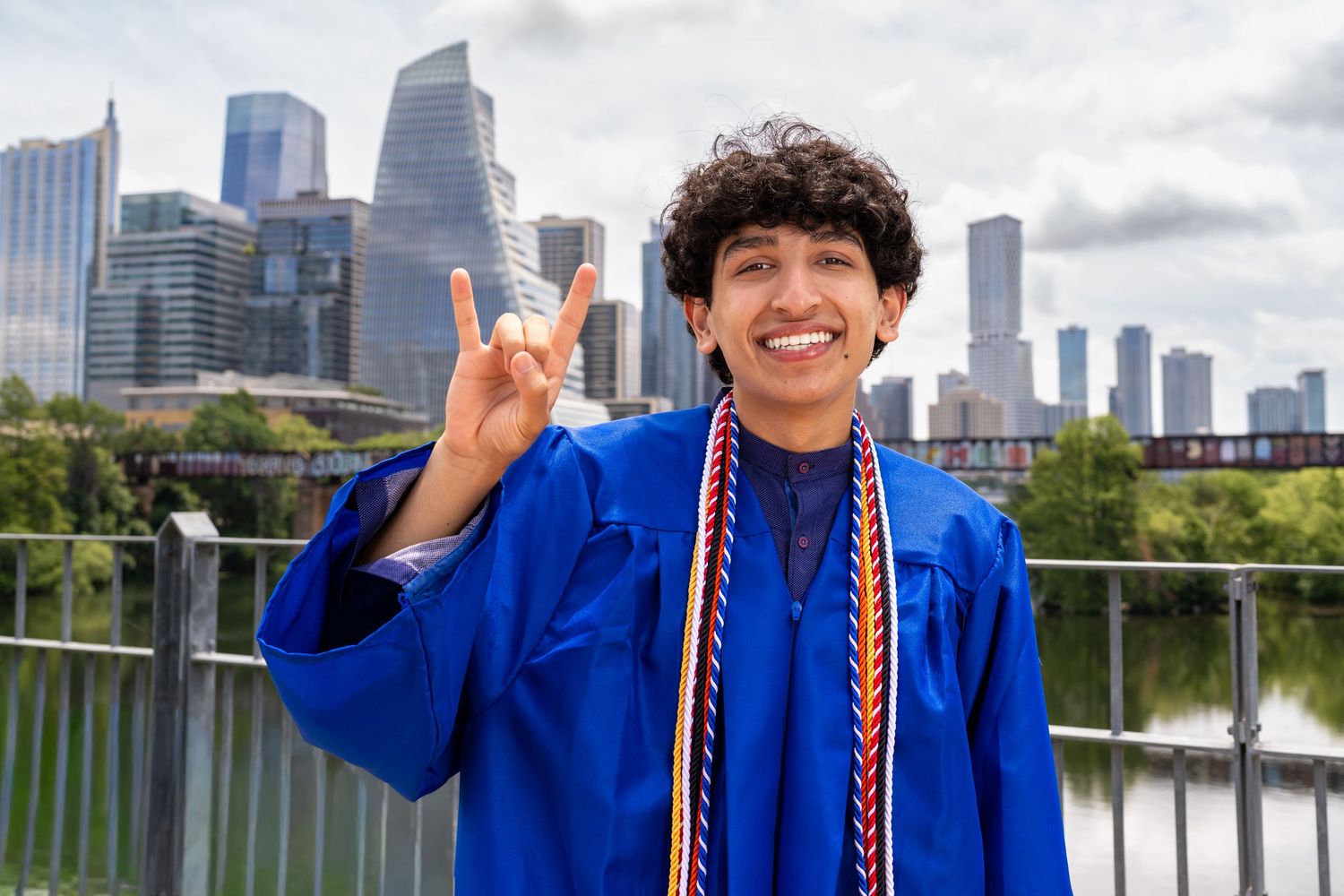 Graduate in blue cap and gown makes hook em horns sign with city skyline backdrop on sunny day.