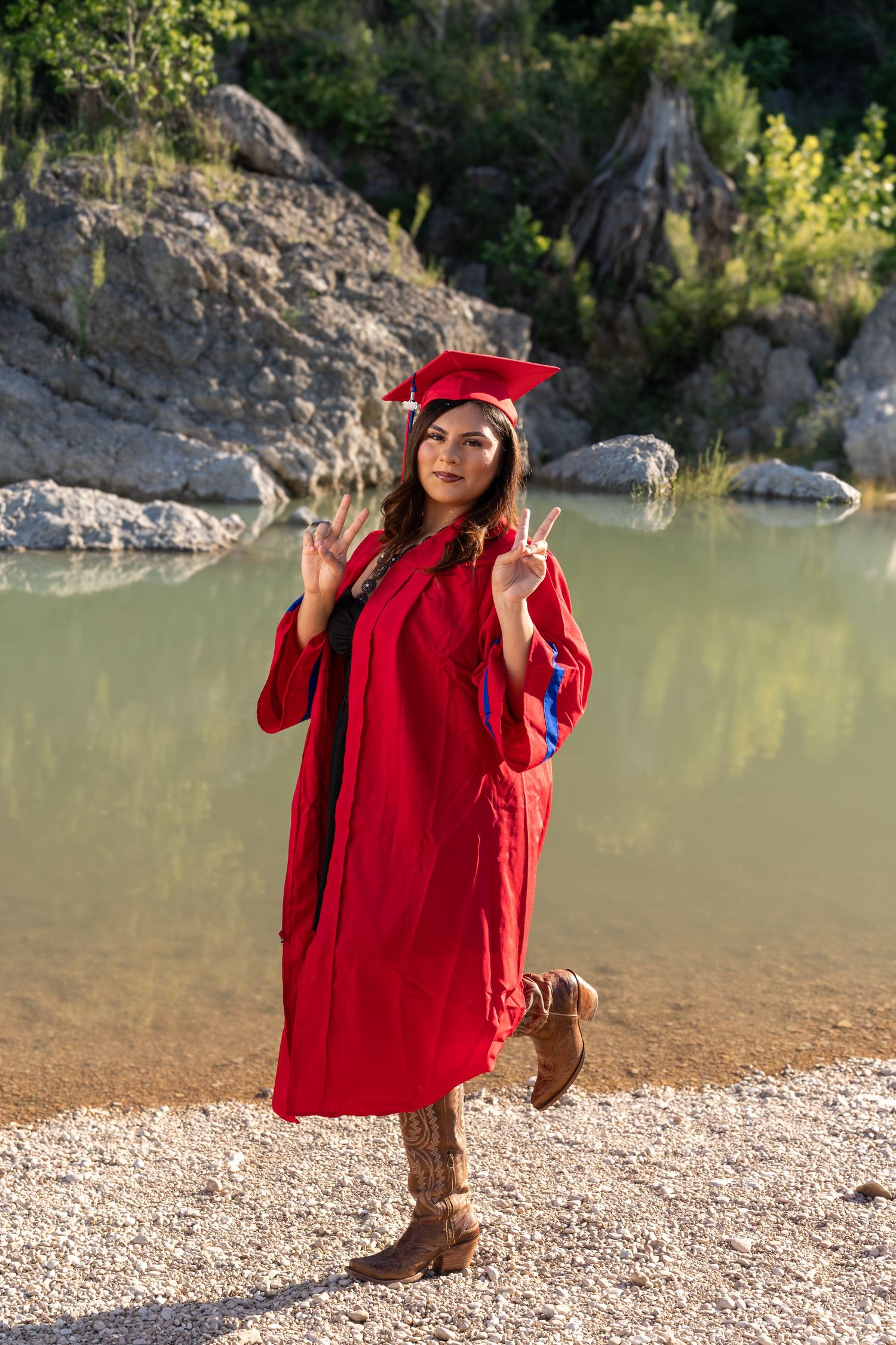 Young woman in red graduation cap and gown poses by a scenic lake with rocks and trees in background.
