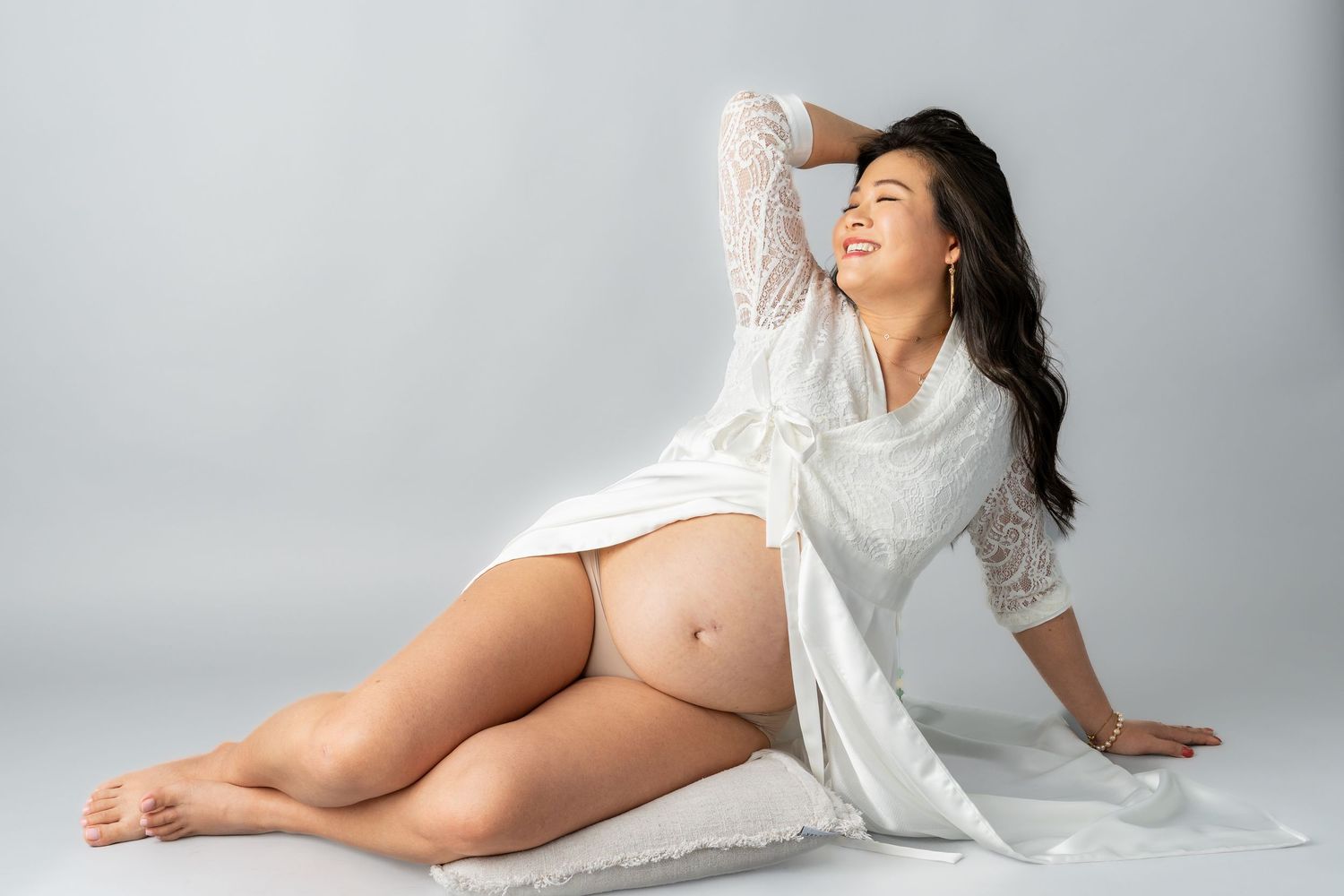 Pregnant woman in white lace robe sitting gracefully on floor, showcasing baby bump in a professional maternity photo shoot.