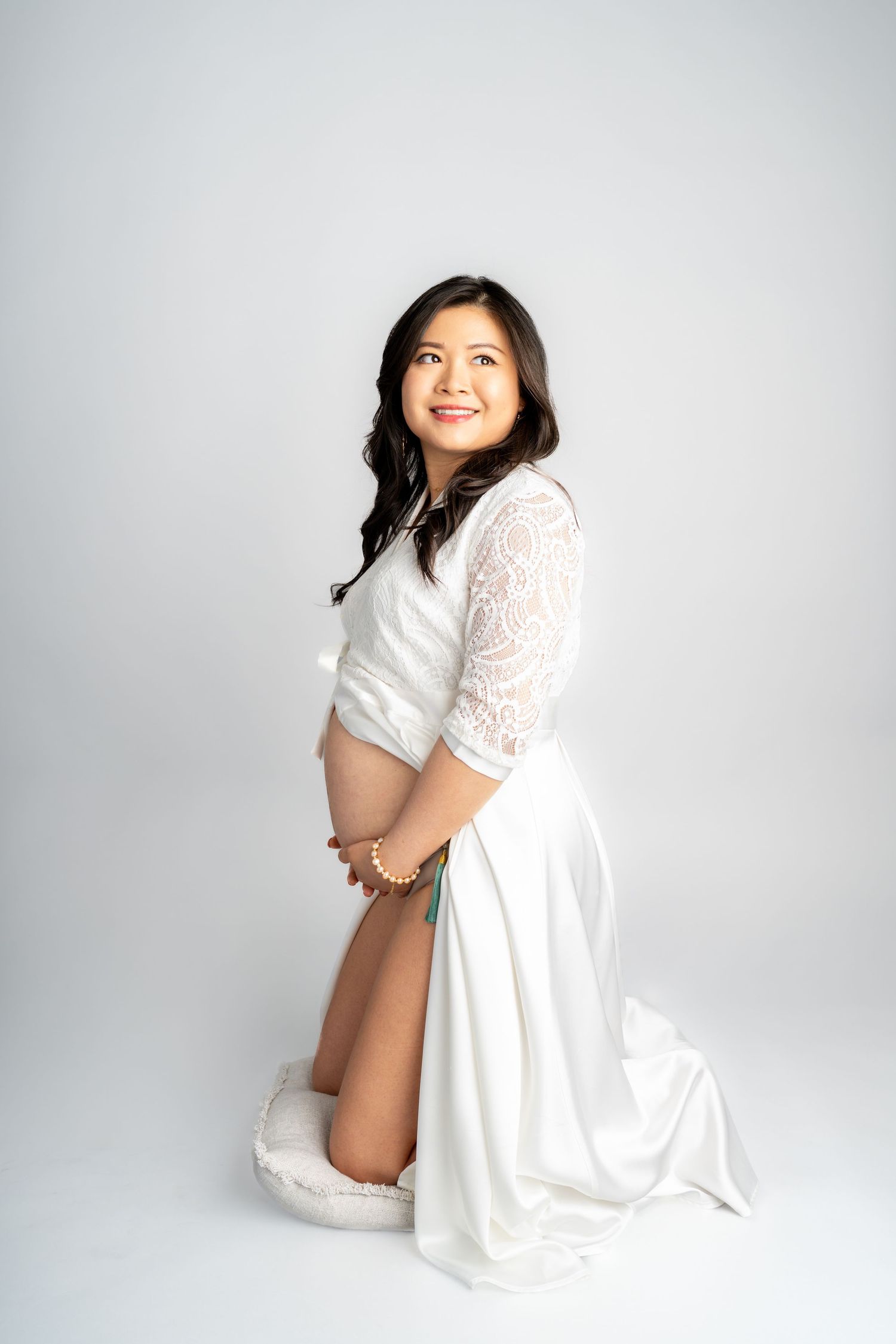 Pregnant woman in white lace gown kneeling gracefully, cradling her baby bump, smiling warmly in a studio setting.