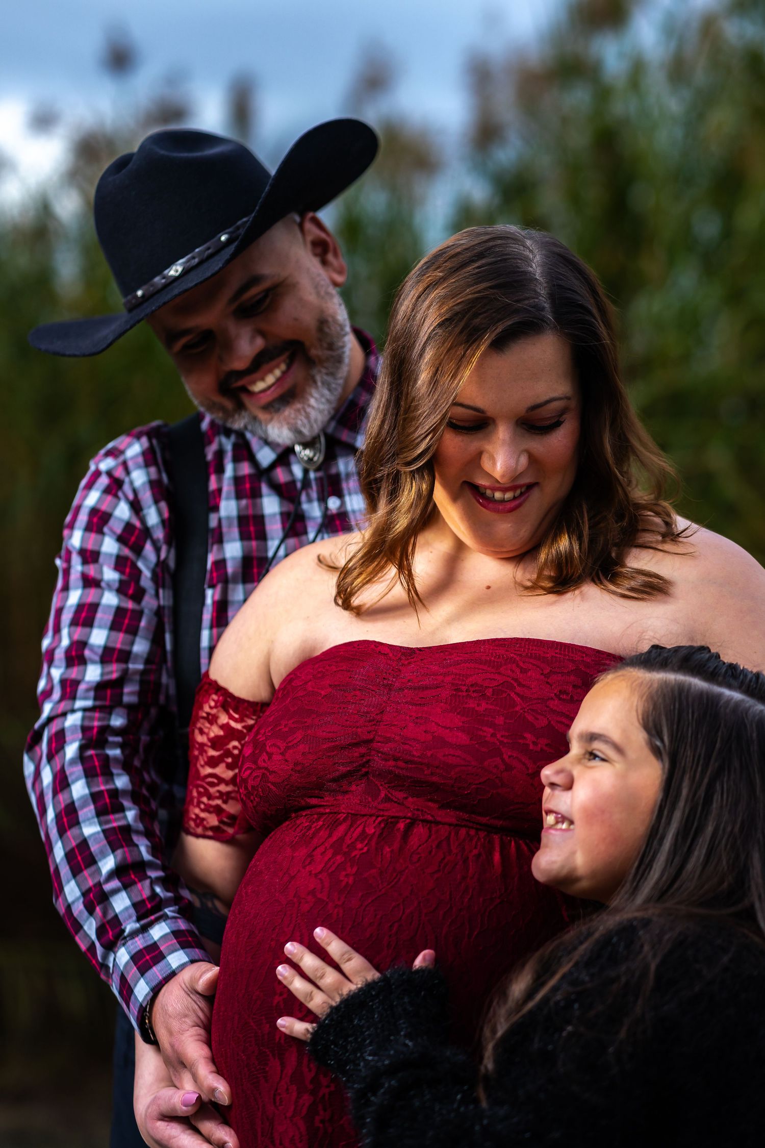 A couple embraces during an outdoor maternity photoshoot, with the woman wearing a burgundy lace dress.