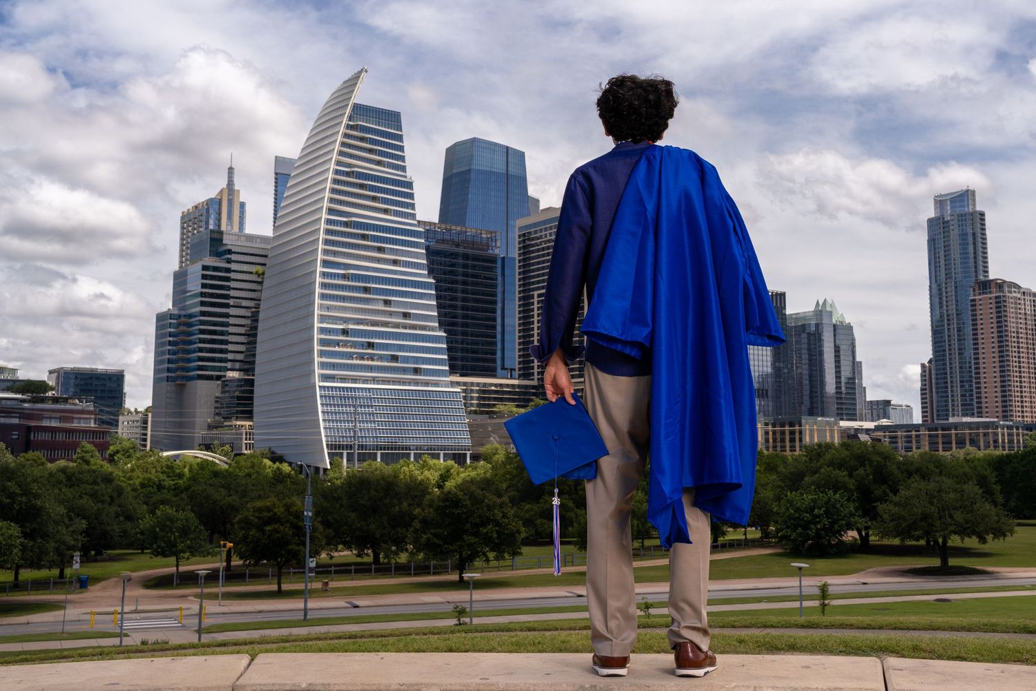 Graduate in blue cap and gown stands overlooking Austin Texas city skyline on cloudy day.