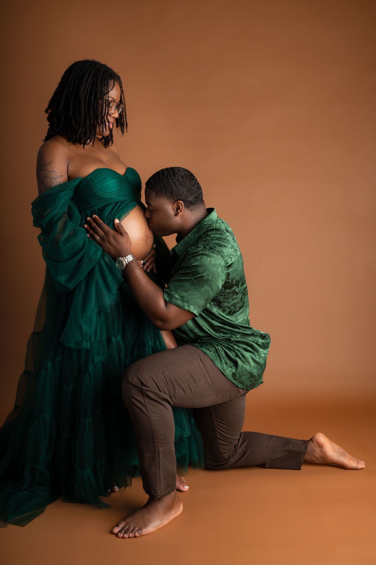 A man kneels kissing a pregnant woman's belly during a maternity photoshoot, both wearing elegant green outfits.