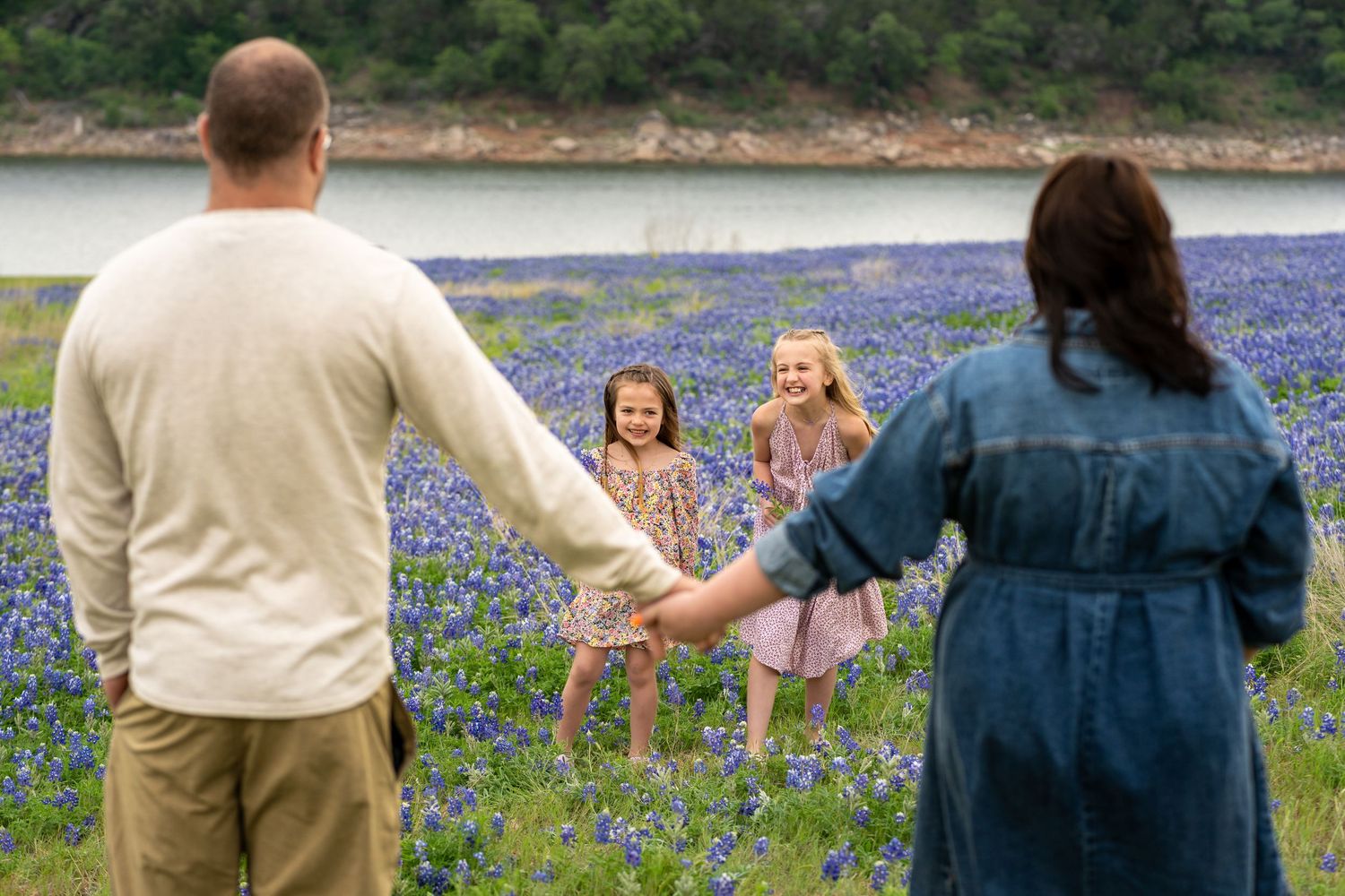 Family holding hands in a bluebonnet field near a lake, with two young girls smiling in matching floral dresses.