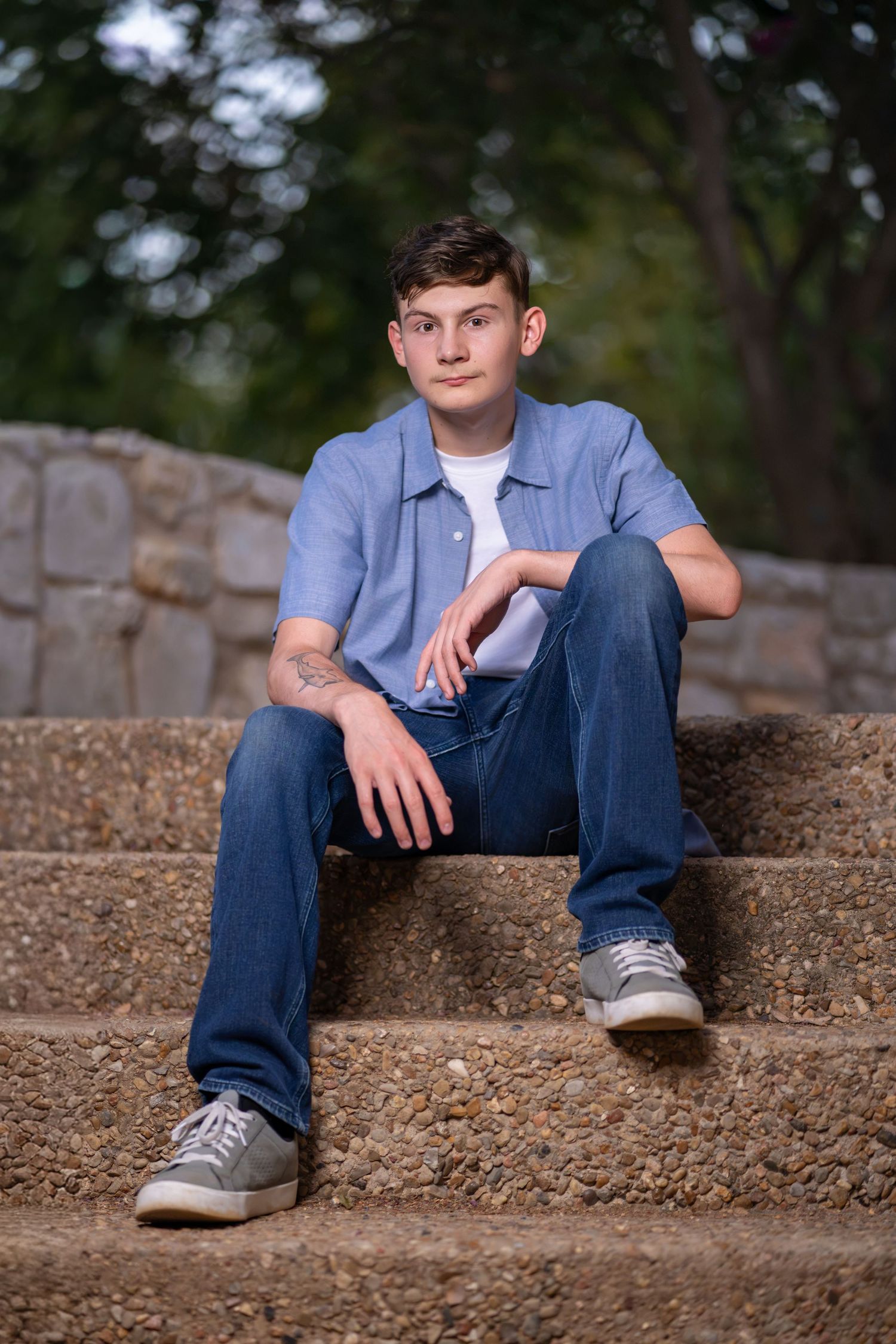 Young man in blue shirt and jeans sitting relaxed on stone steps outdoors with trees in background.