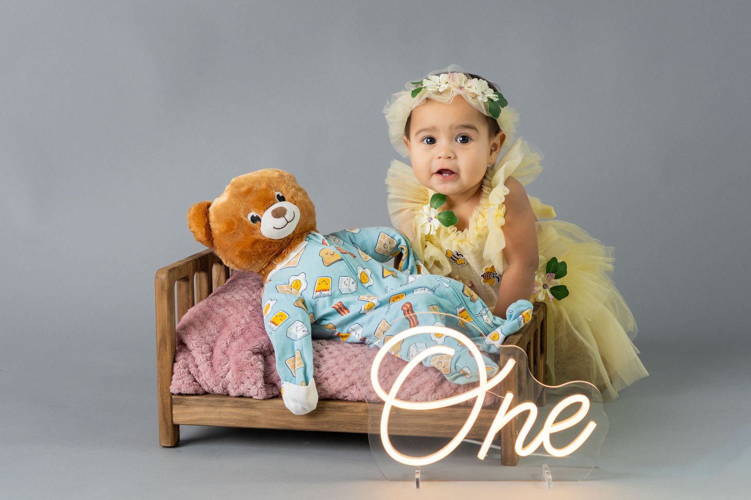 Baby girl in yellow tutu and floral crown sits on mini wooden bed with teddy bear beside a glowing One neon sign.