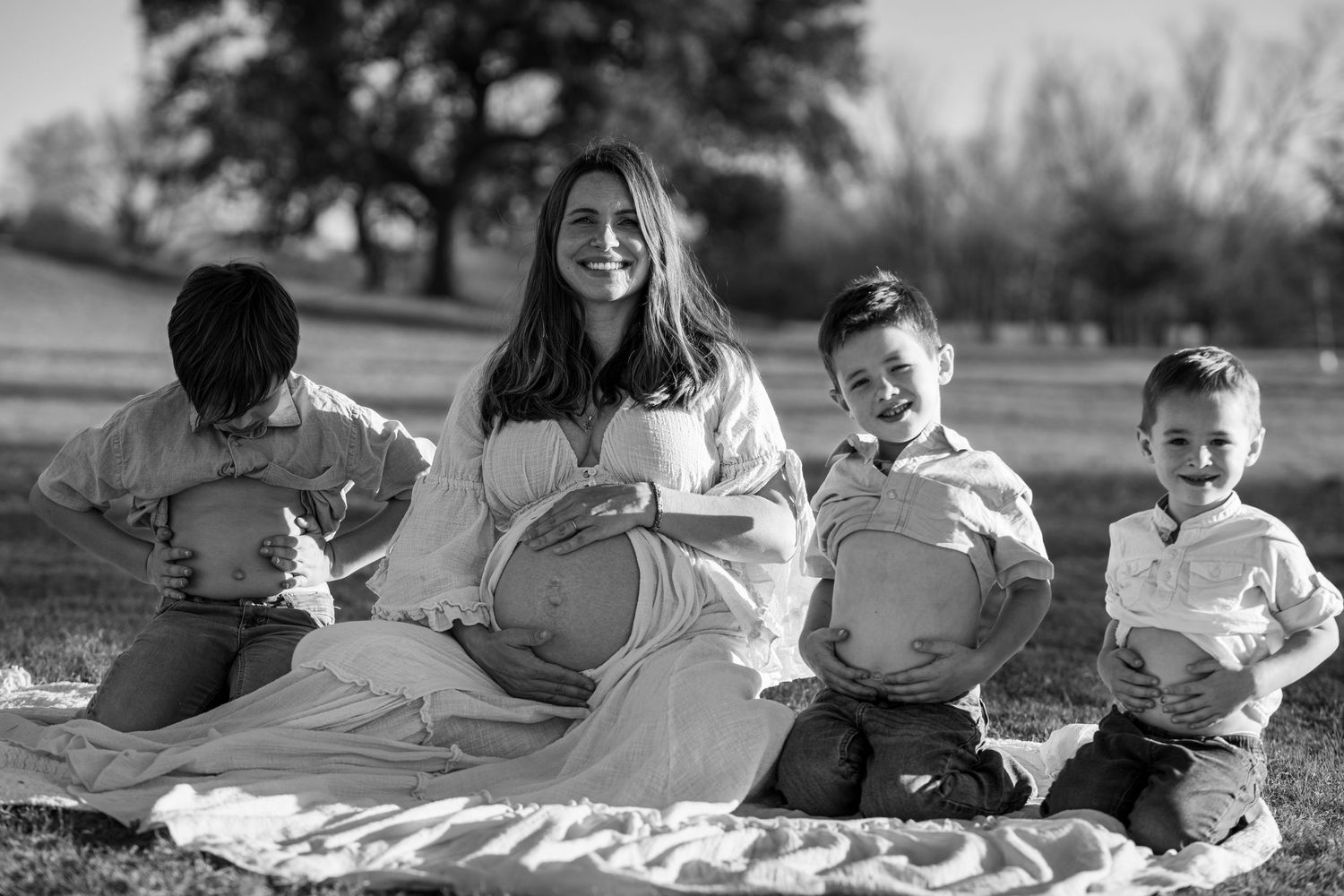Black and white photo of a smiling pregnant woman sitting outdoors with three young boys mimicking her baby bump pose.