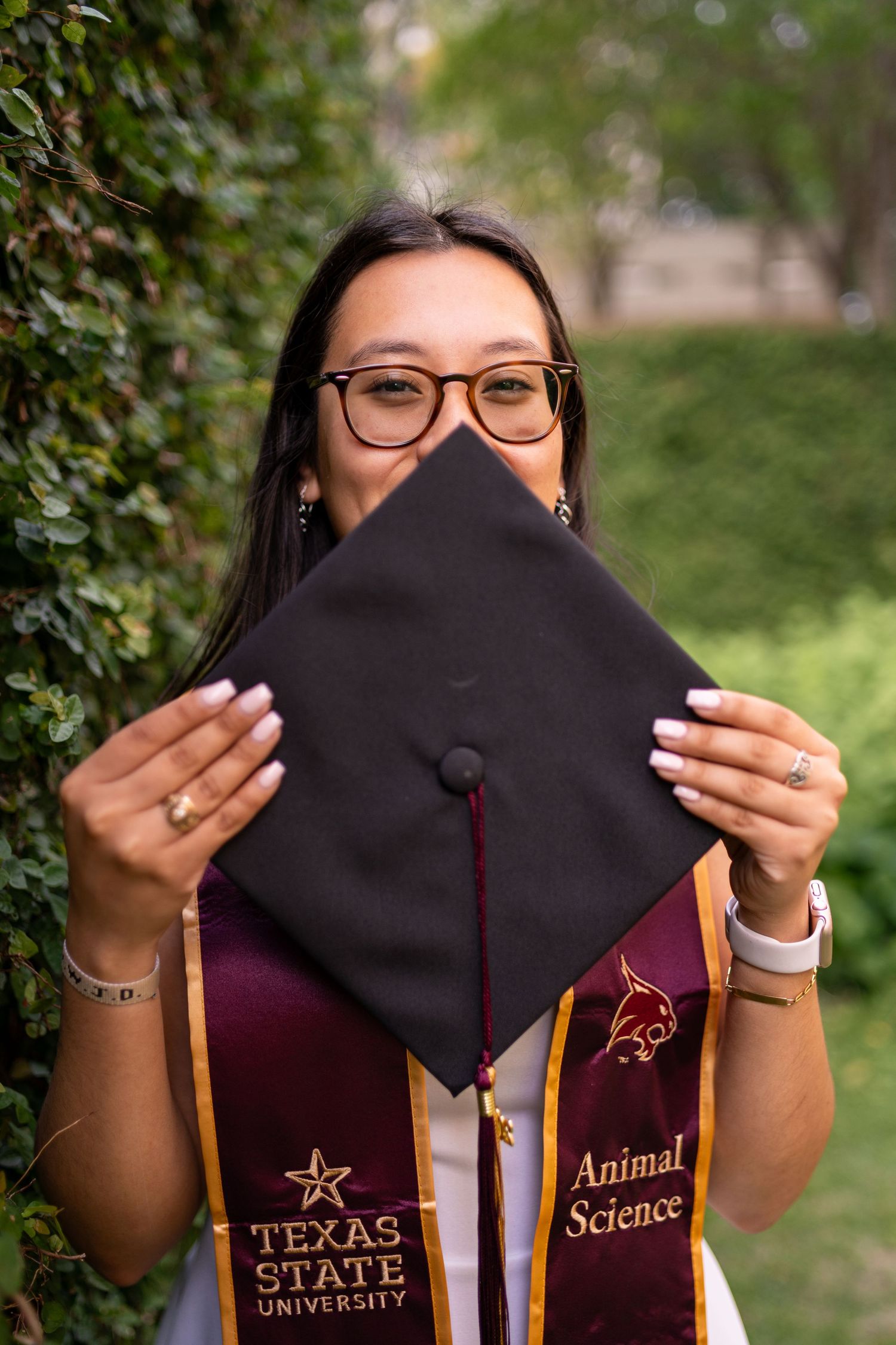 Texas State University Animal Science graduate holds black mortarboard cap outdoors wearing maroon graduation stole and glasses.