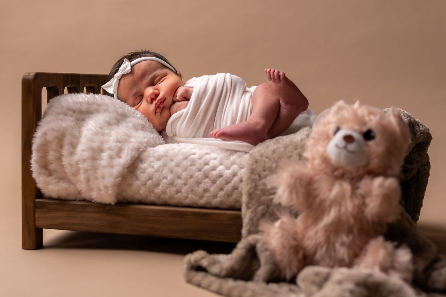 Newborn baby sleeping peacefully on tiny wooden bed next to plush teddy bear in warm studio lighting.