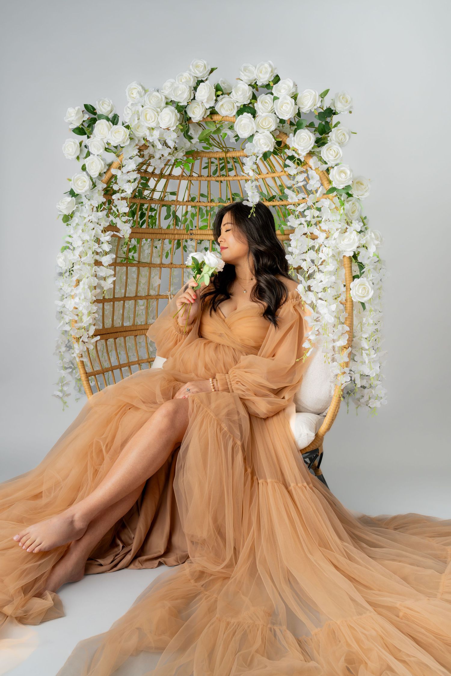 A woman in a flowing champagne tulle gown sits in a floral rattan egg chair with white roses in a studio.