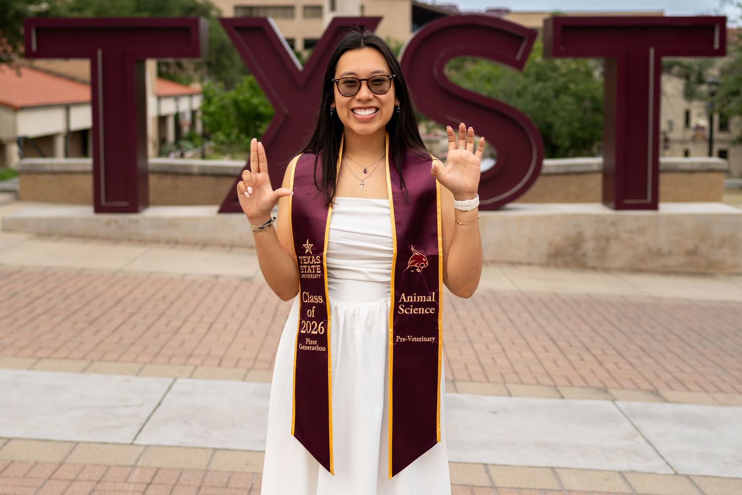 Texas State University graduate poses proudly in maroon stole and sunglasses in front of TXST letters on campus.