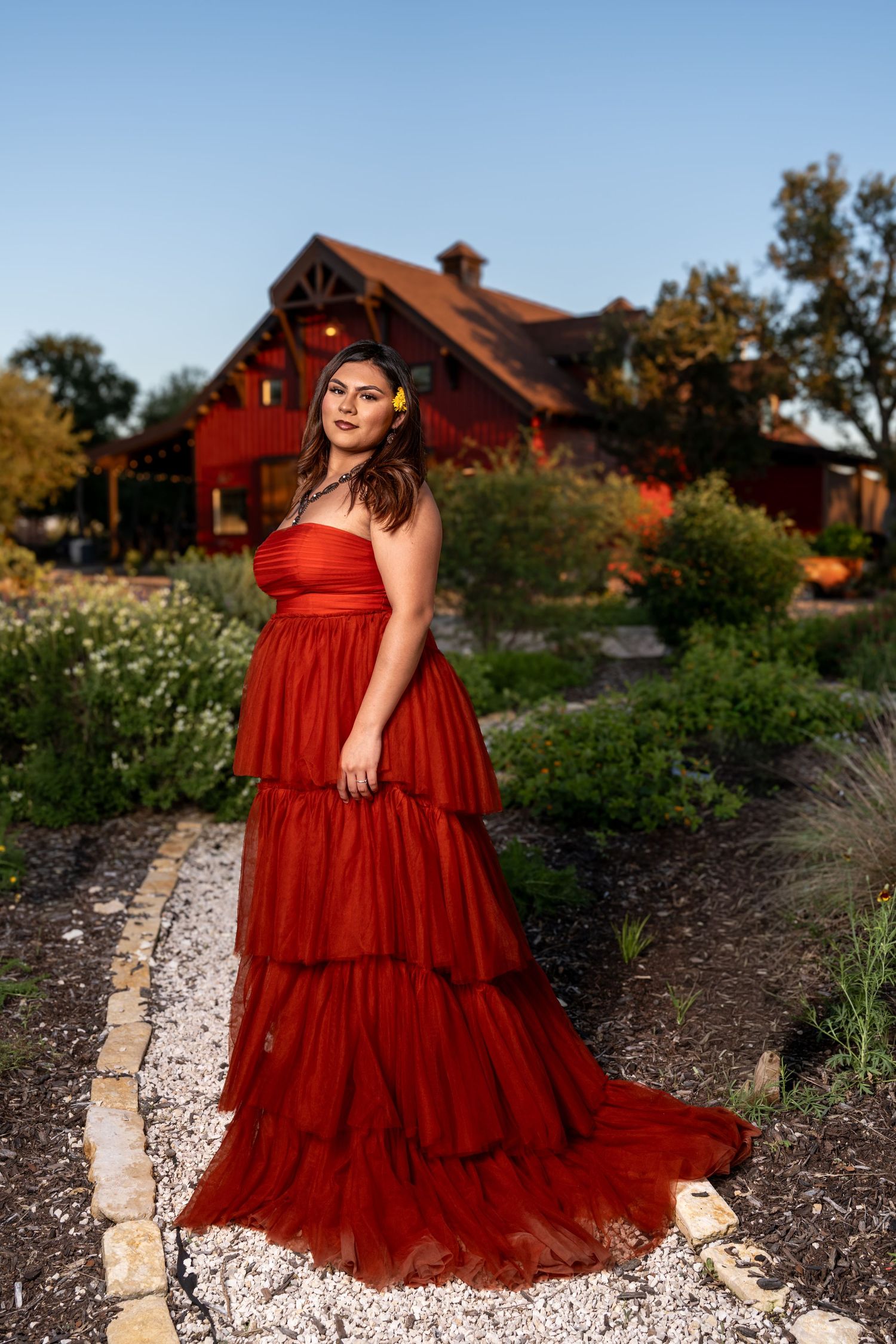Woman in red tiered gown with yellow flower hair accessory poses in garden path before rustic red barn at dusk.