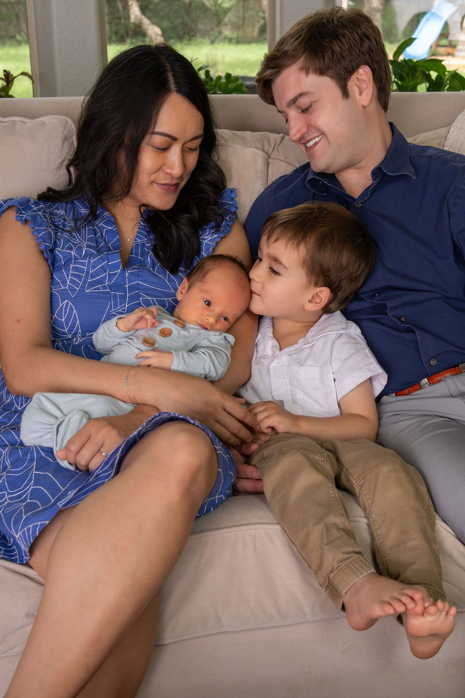 Family of four sits on couch, mom holds newborn while toddler leans in for a kiss, dad smiles warmly.