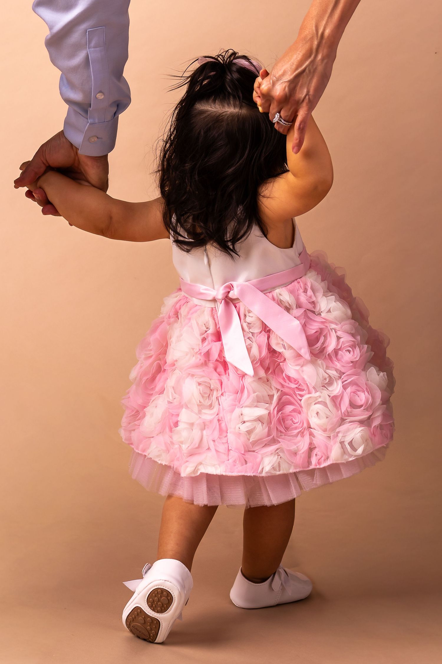A small child in a pink tulle dress with rosettes holds hands with an adult while learning to walk.