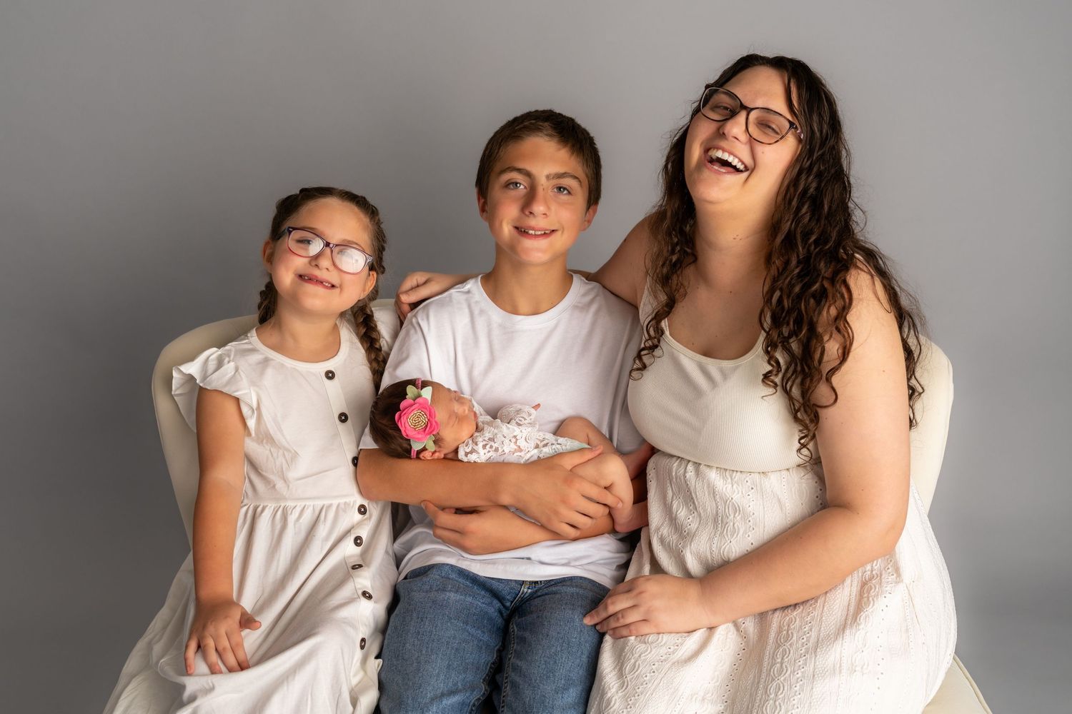 A woman and two children in white outfits pose with a newborn baby in a studio portrait setting.