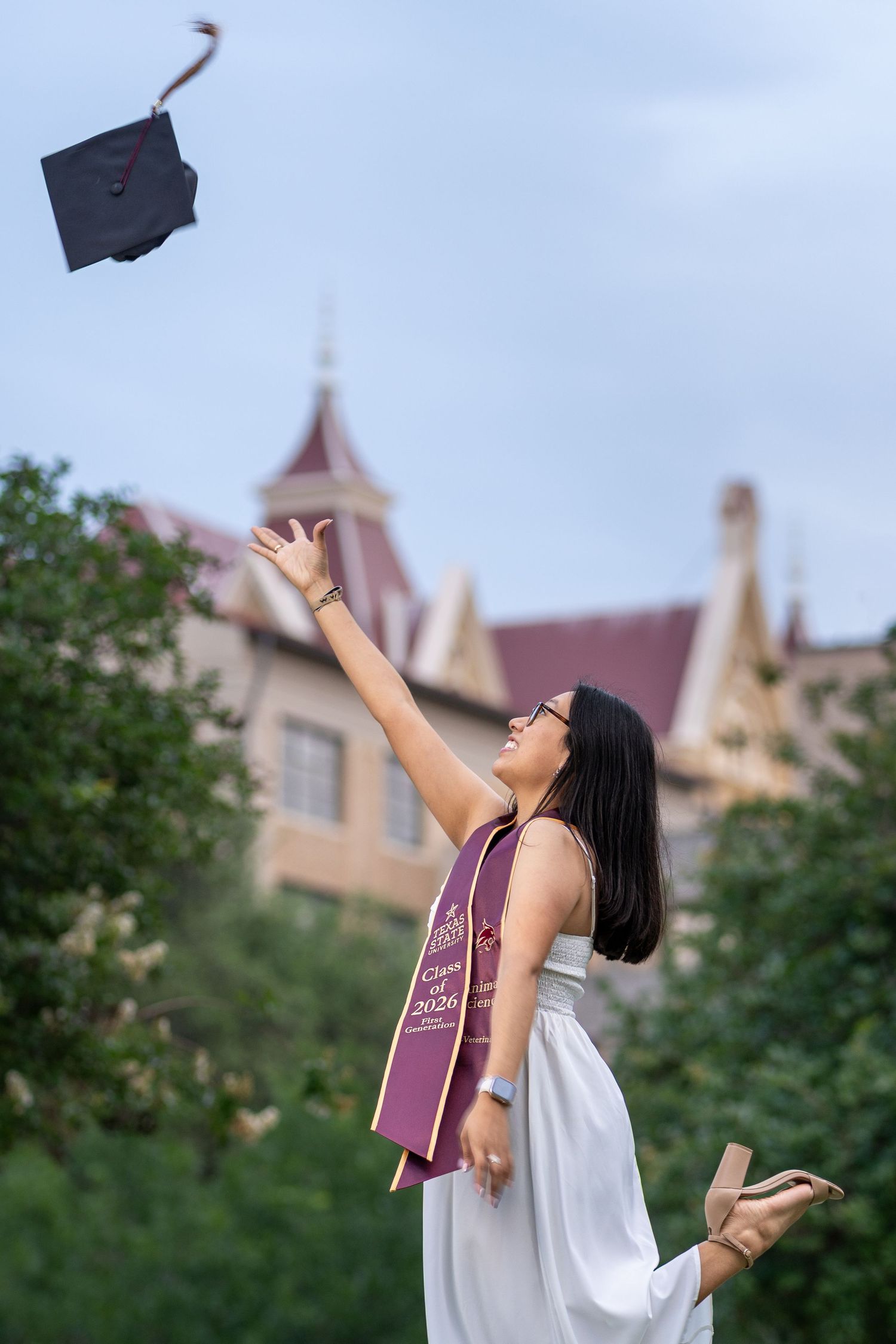Graduate in white dress tossing mortarboard cap in the air outdoors near a historic university building.