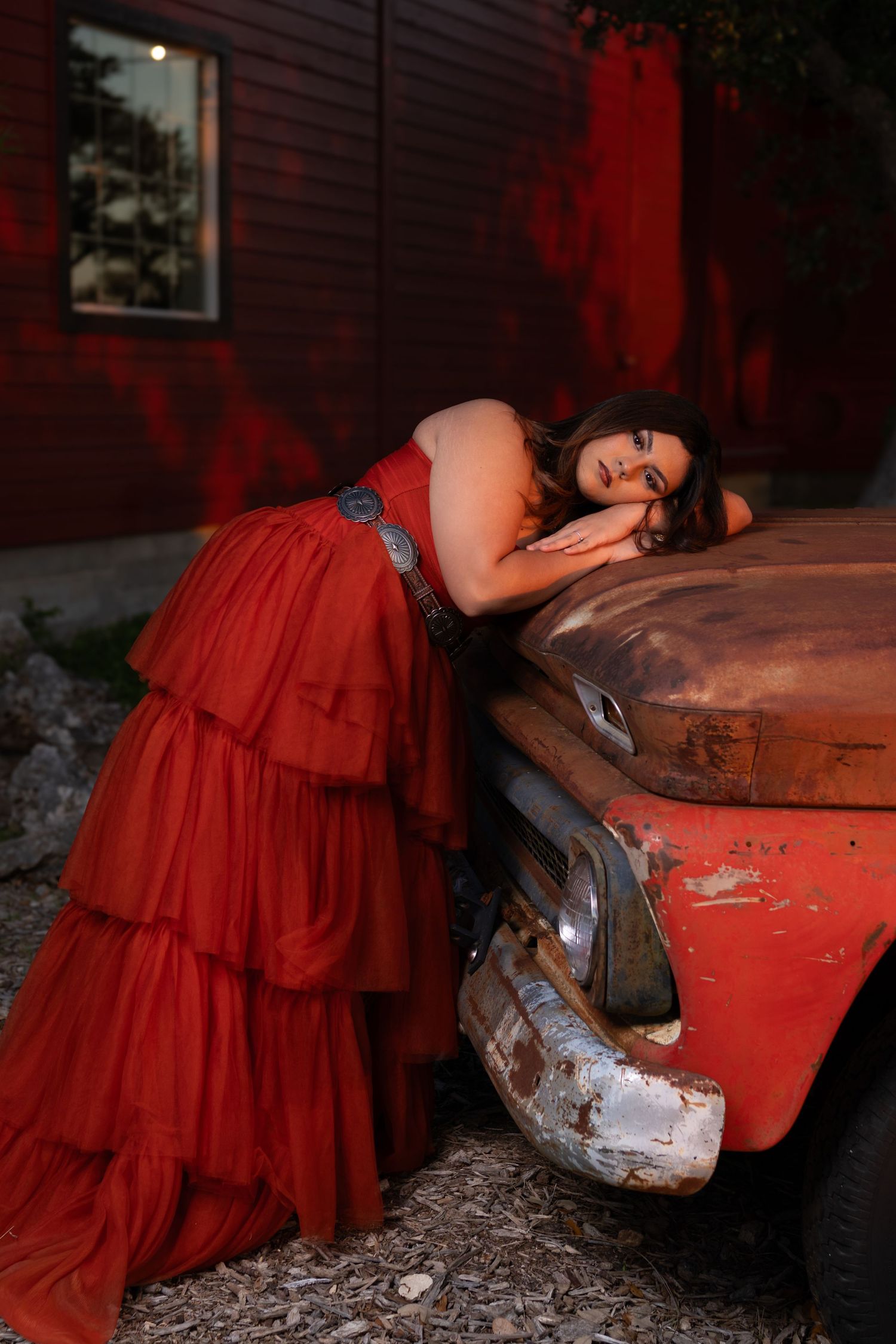 Woman in red ruffled gown leans gracefully on a rusty vintage car near a red barn at dusk.