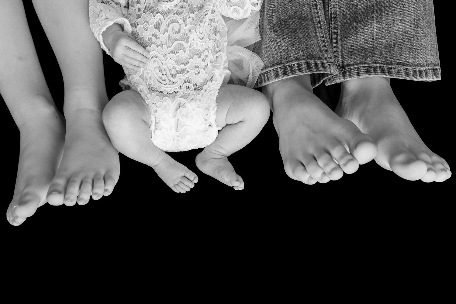 Black and white photo of three generations of bare feet, including a baby, child, and adult, dangling against a dark background.