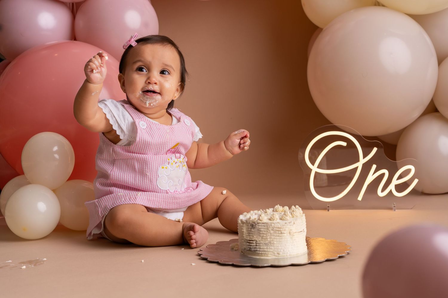 Baby in pink dress reaches for birthday cake during cake smash photo session with balloon decorations.