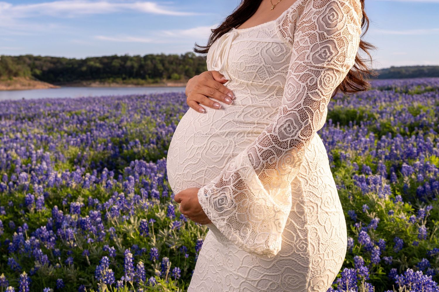 Pregnant woman in white lace dress cradling baby bump in a vibrant purple wildflower field under a blue sky.