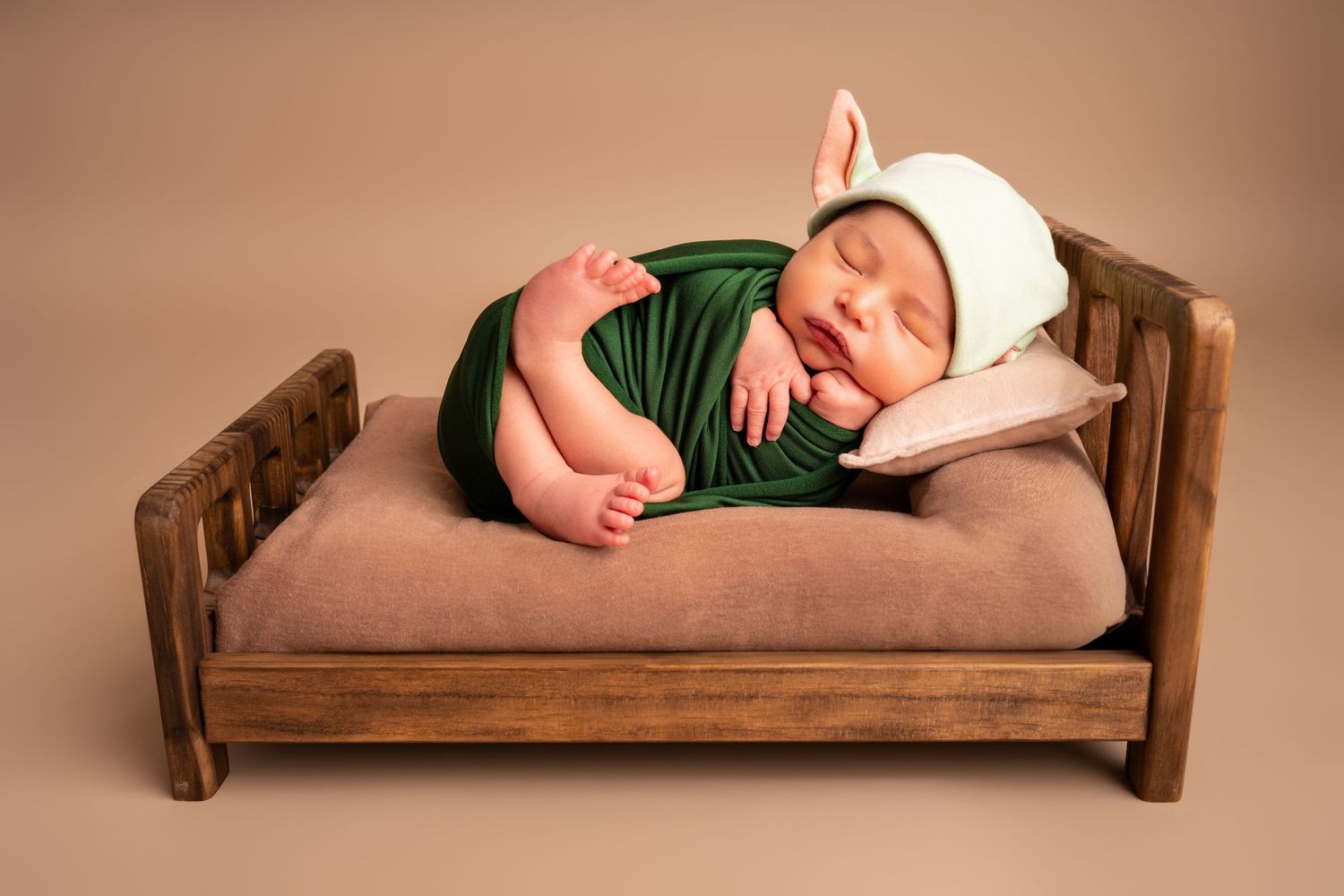 Sleeping newborn baby in a green wrap and white pointed-ear hat resting on a small wooden bed with a tan mattress and pillow.