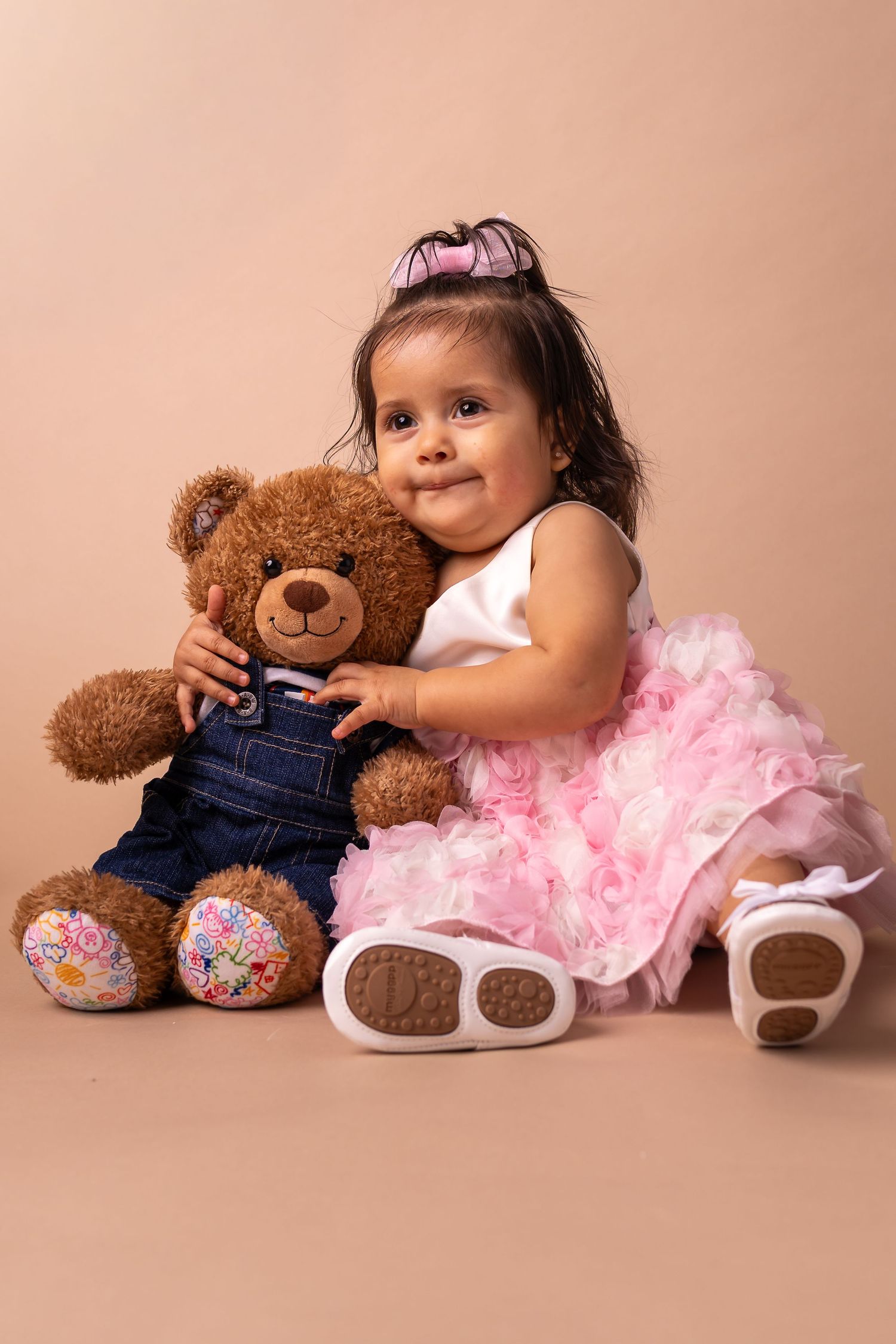 Smiling baby girl in pink tutu dress hugging a brown teddy bear in denim overalls on a beige background.