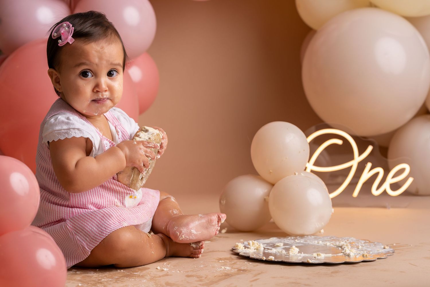 Baby girl in pink romper smashing first birthday cake surrounded by pink and cream balloons with One sign.